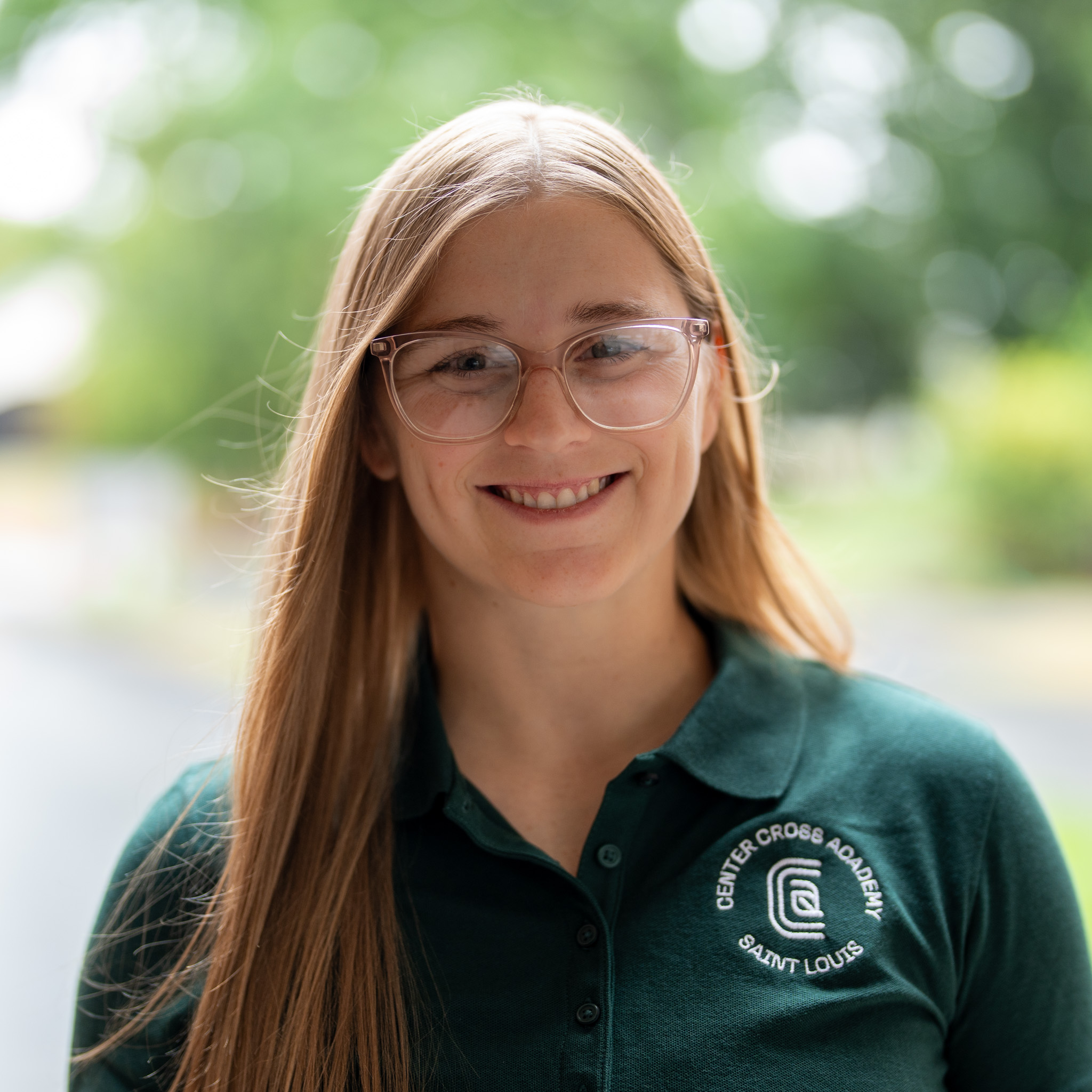 A woman with long blonde hair and glasses smiles outdoors, wearing a dark green polo shirt with a Center Cross Academy Saint Louis logo. The background is blurred greenery.