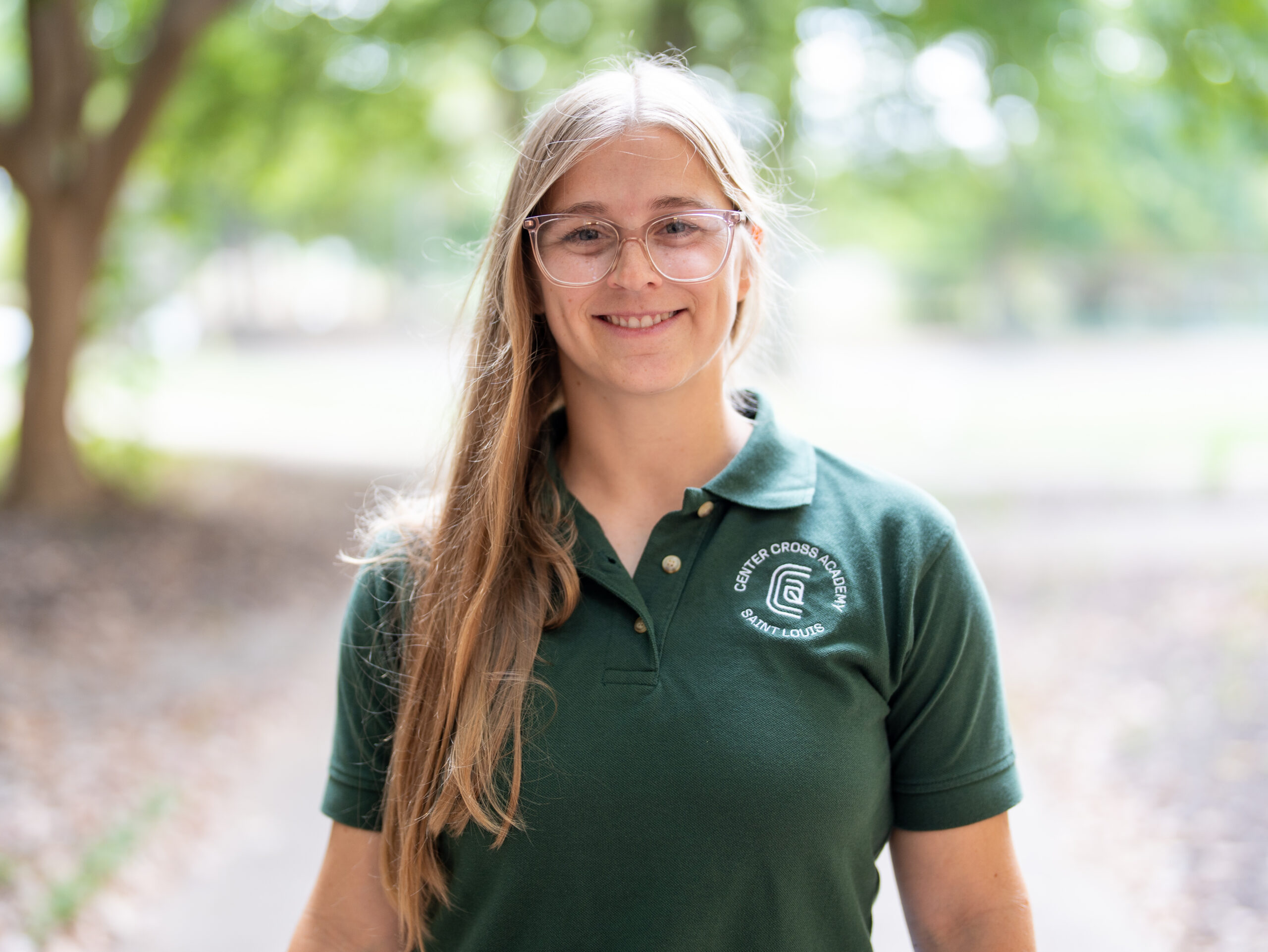 A woman with long blond hair and glasses smiles outdoors, wearing a green polo shirt with a white circular logo that reads Centre for Good Food Gardening. Trees and soft sunlight are in the background.