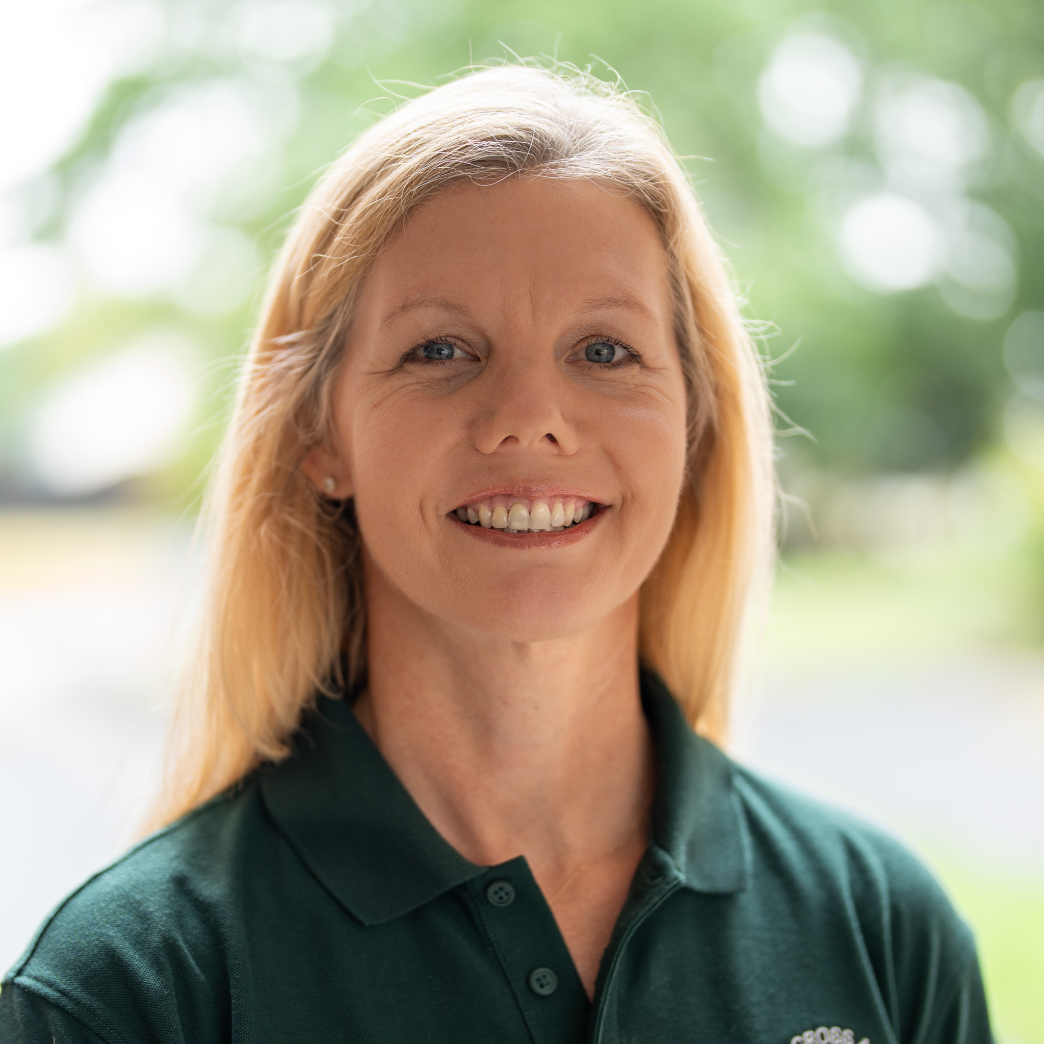 A woman with long blonde hair, wearing a dark green collared shirt, smiles at the camera. She is outdoors with blurred trees and greenery in the background.