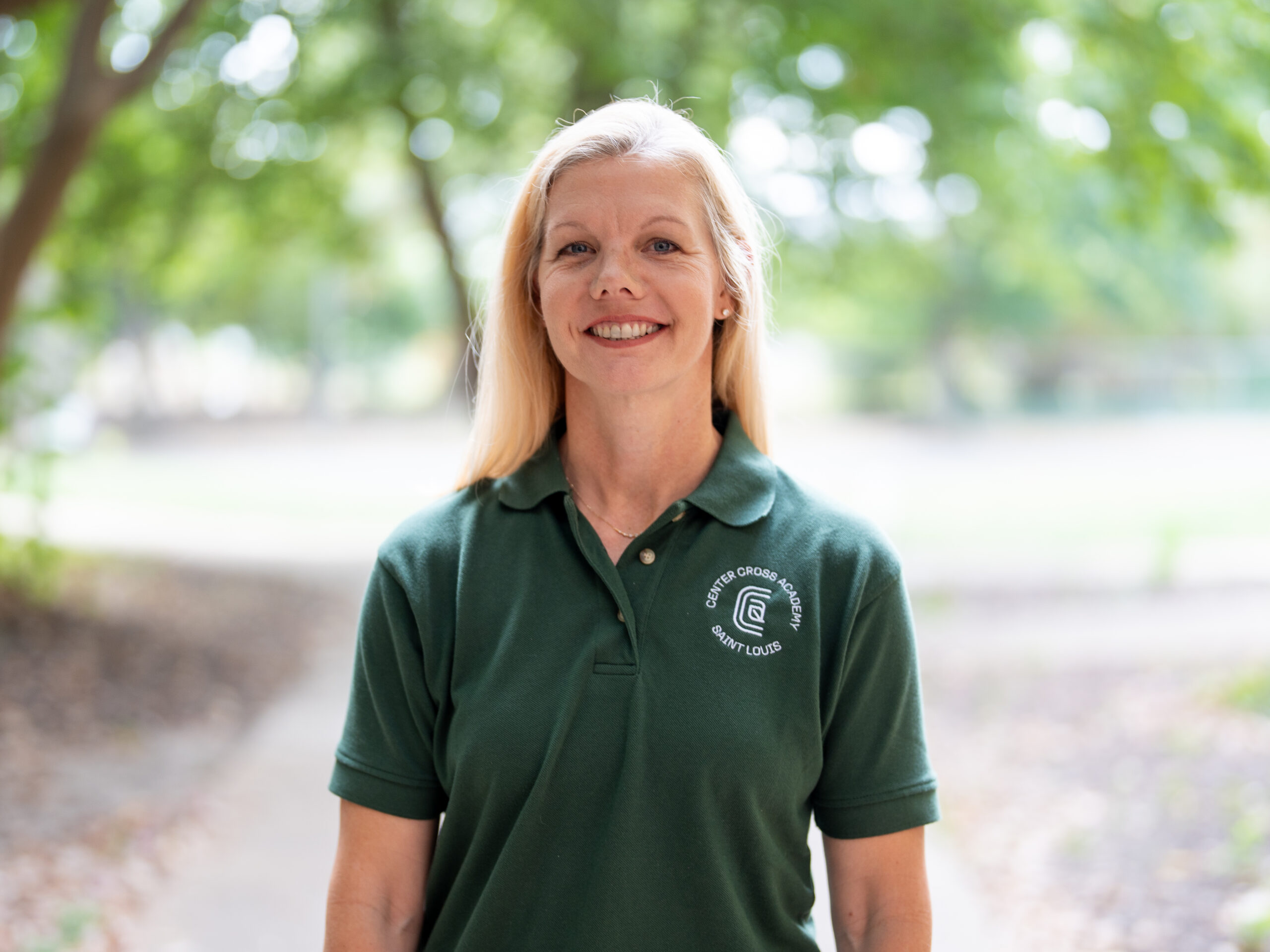 A woman with long blonde hair wearing a dark green polo shirt with a logo on the left chest stands outdoors, smiling, with trees and a pathway in the blurred background.
