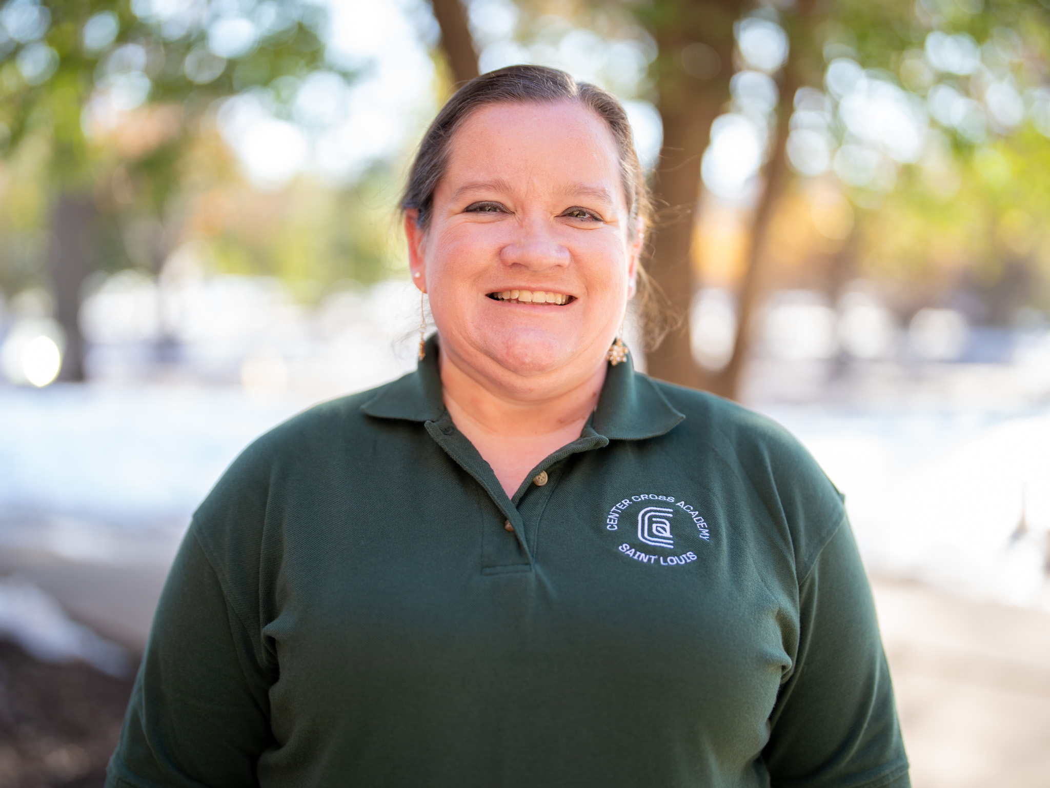 A woman smiles outdoors while wearing a dark green polo shirt with a circular logo that reads Saint Charles Parks, Saint Louis. Trees and sunlight are visible in the blurred background.