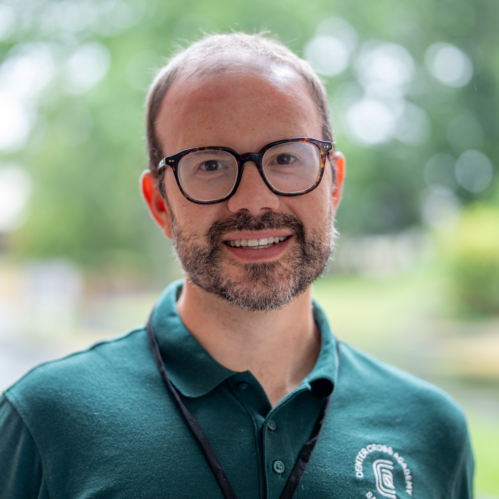 A man with glasses and a beard, wearing a green polo shirt and lanyard, smiles outdoors with trees and soft daylight in the blurred background.