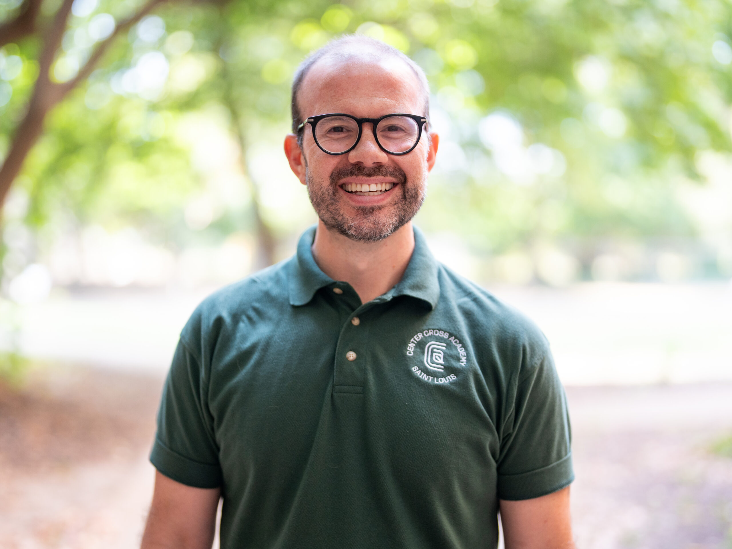 A smiling man with glasses and a beard stands outdoors. He is wearing a green polo shirt with a white logo and text on the left side. The background is blurred trees and natural light.