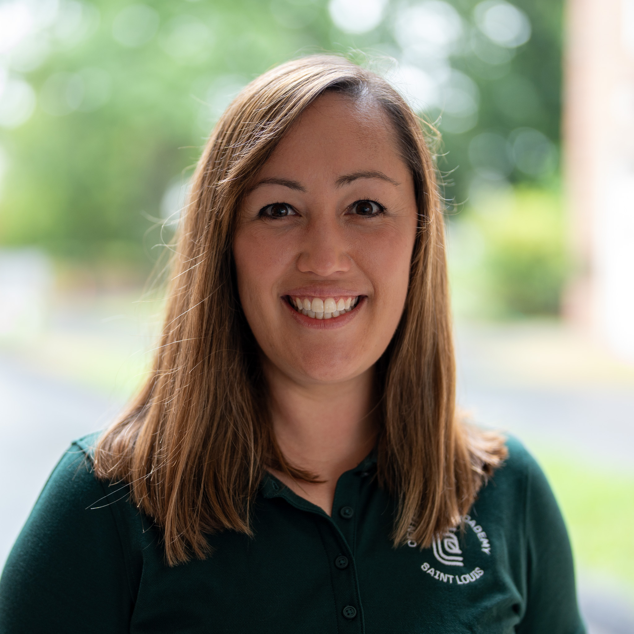A woman with straight, light brown hair smiles at the camera. She is wearing a dark green collared shirt and standing outdoors with blurred greenery in the background.