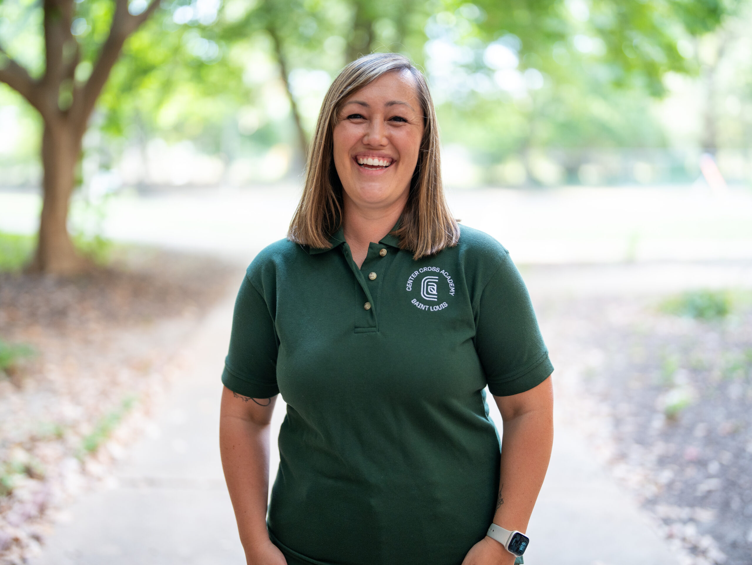 A woman with straight, light brown hair stands outdoors on a walkway, smiling. She wears a dark green polo shirt with a white logo and a watch. Trees and sunlight are visible in the blurred background.