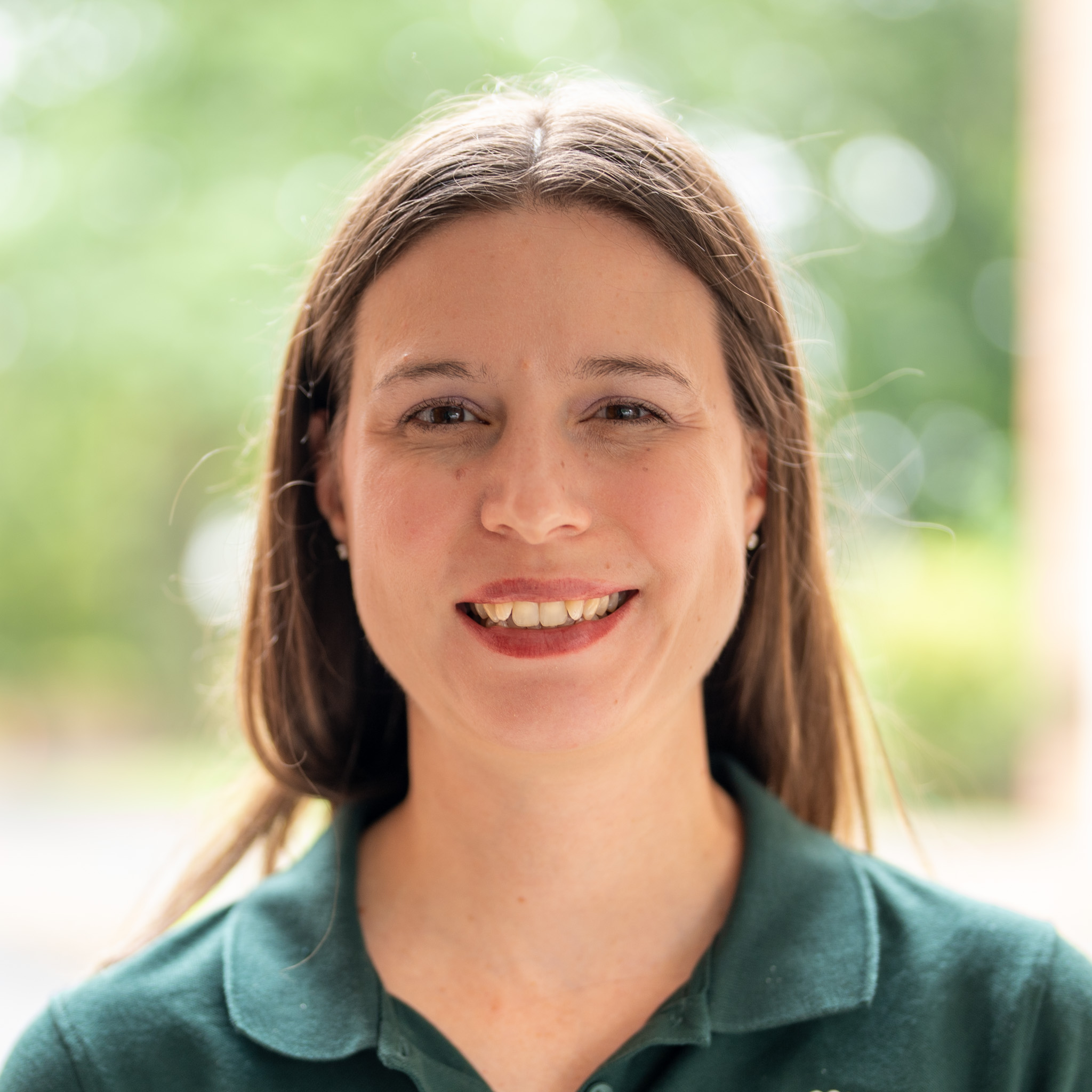 A woman with straight brown hair and a green collared shirt smiles at the camera, standing outdoors with a blurred green background.