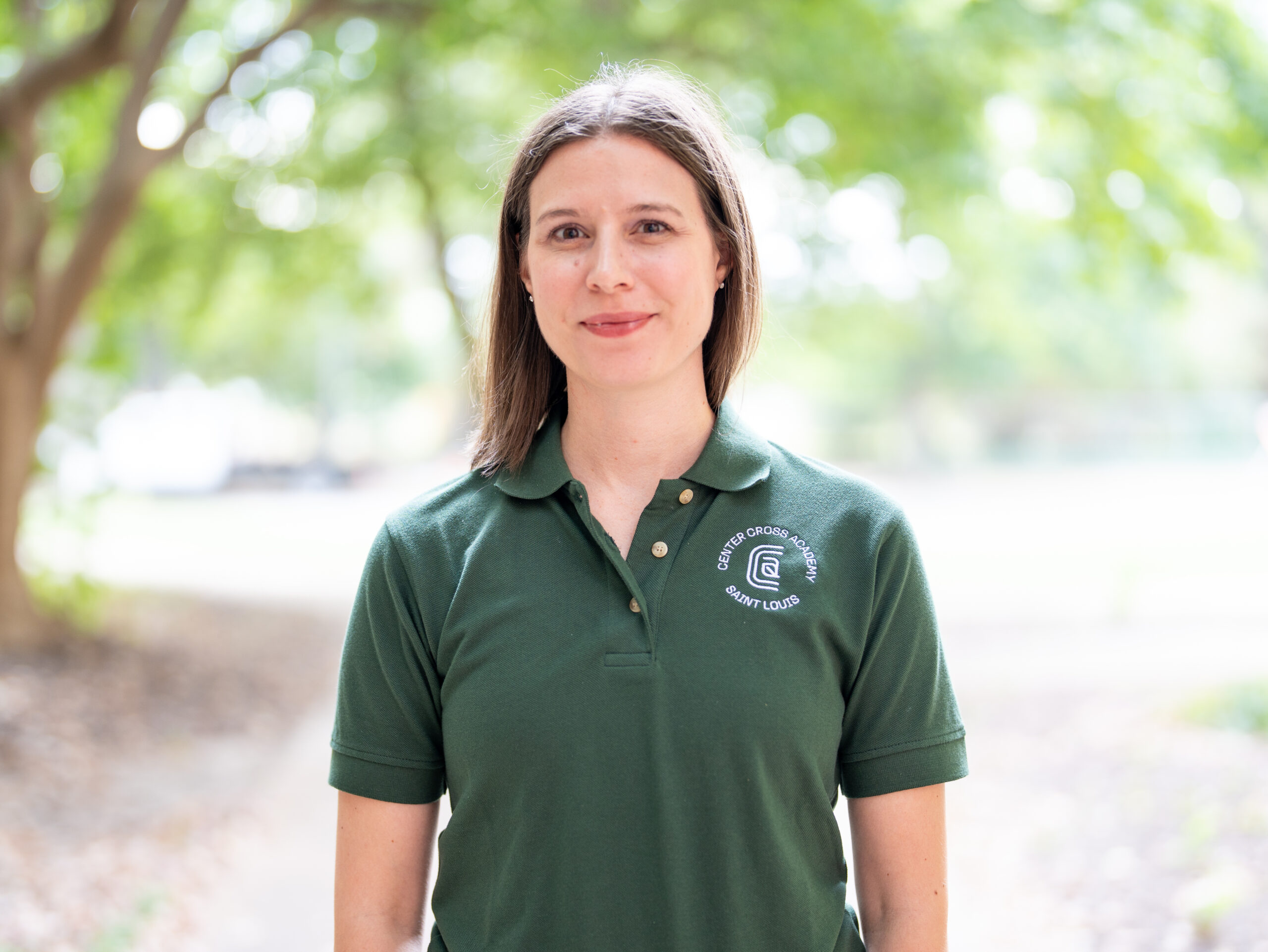 A woman with straight brown hair stands outdoors, wearing a dark green collared shirt with a white logo. She smiles softly, and leafy trees blur in the background.