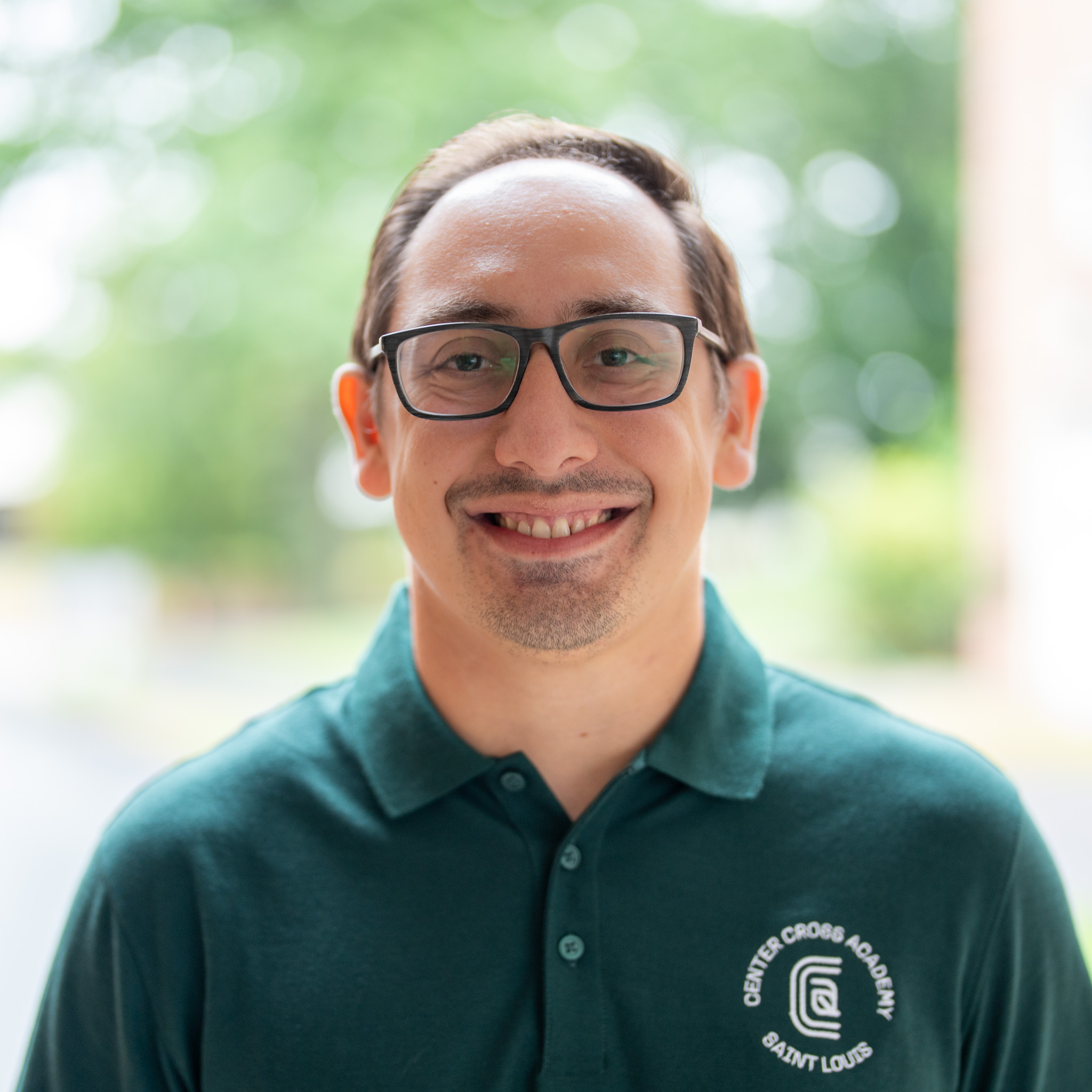 Smiling man with glasses and short brown hair wearing a green polo shirt with a Christ Cross Academy Saint Louis logo, standing outdoors with a blurred green background.