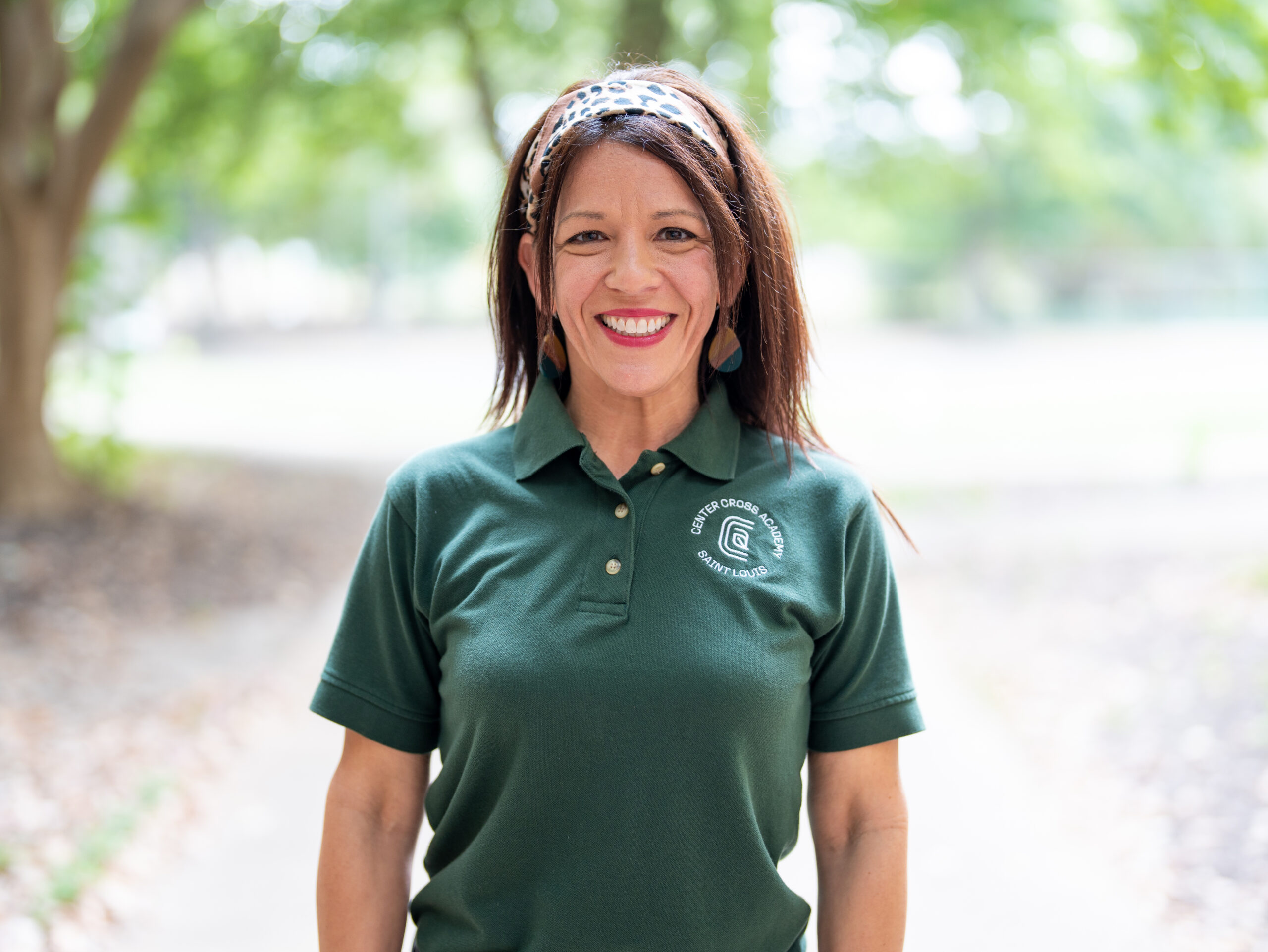 A woman with long brown hair, wearing a patterned headband and a dark green polo shirt with a logo, stands smiling outdoors on a path with trees in the background.