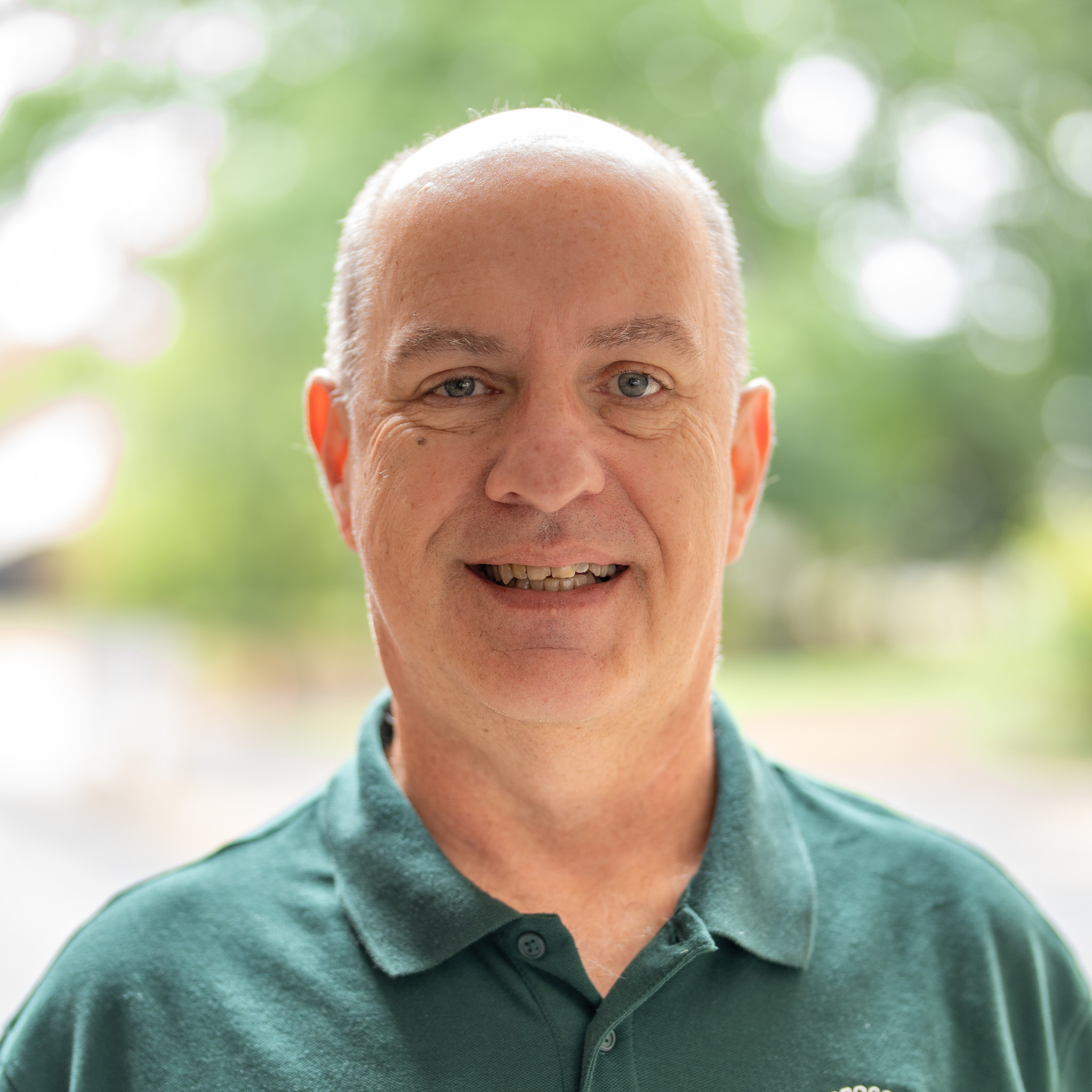 A middle-aged man with short hair and a green polo shirt smiles outdoors with trees and soft sunlight in the background.