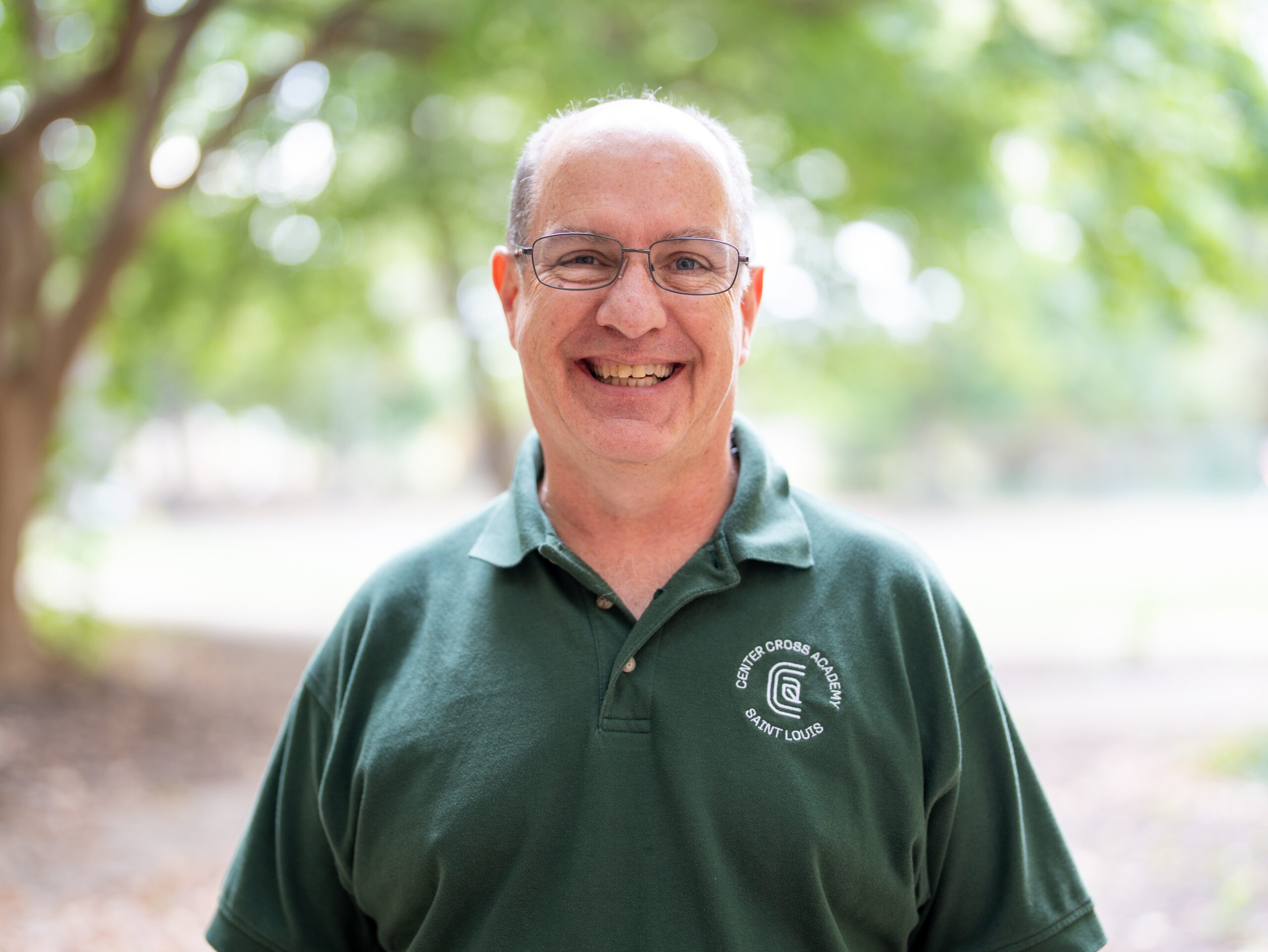 A smiling older man with glasses stands outdoors, wearing a green polo shirt with a white logo. The background is blurred with trees and sunlight.