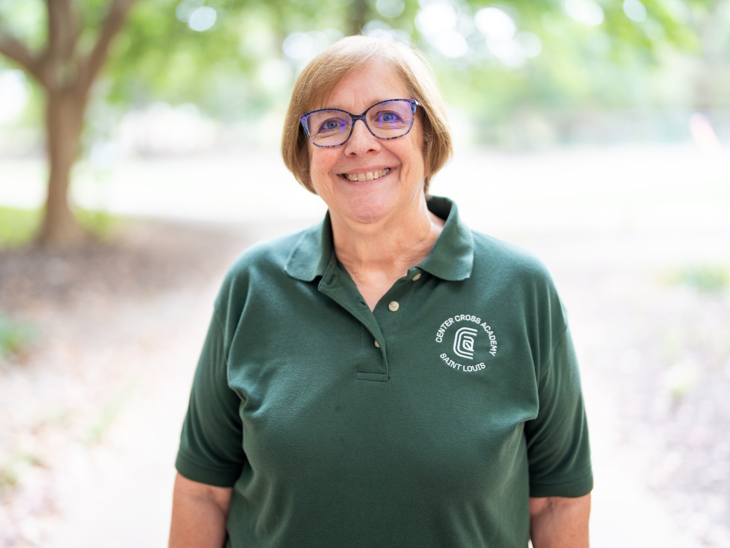 A smiling older woman with short light brown hair and glasses wears a green polo shirt with a “Carondelet Garden & School Saint Louis” logo, standing outdoors with trees and soft natural light in the background.