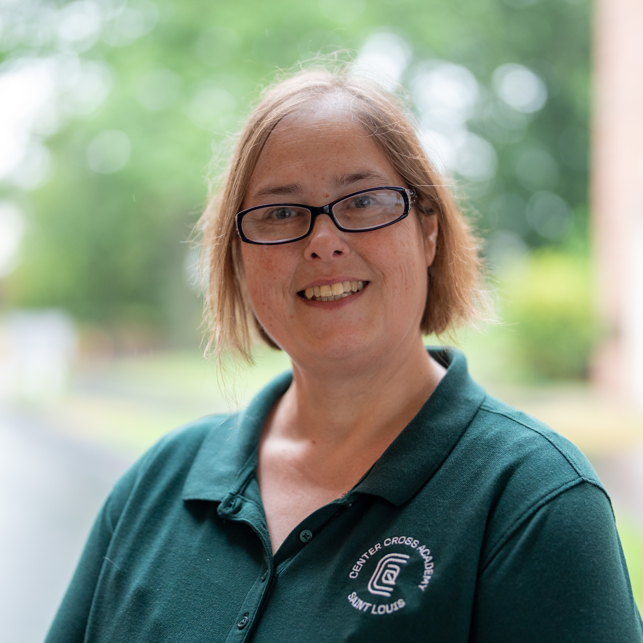 A woman with short light brown hair and glasses smiles outdoors. She wears a dark green collared shirt with a Center Cross School Saint Louis logo. The background is blurred greenery and a path.