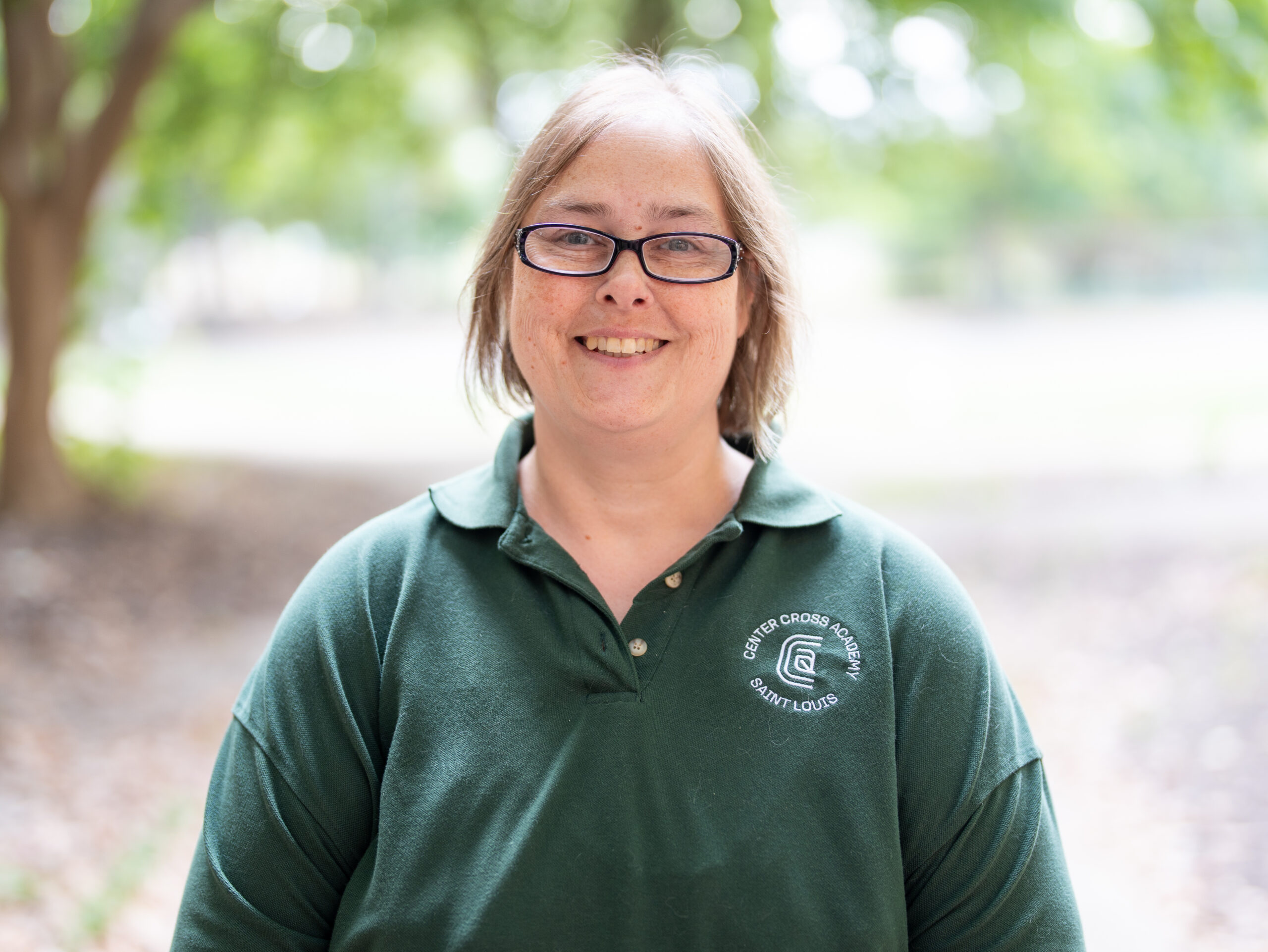 A smiling person with glasses, wearing a green collared shirt with a white logo, stands outdoors in a blurred, leafy setting.