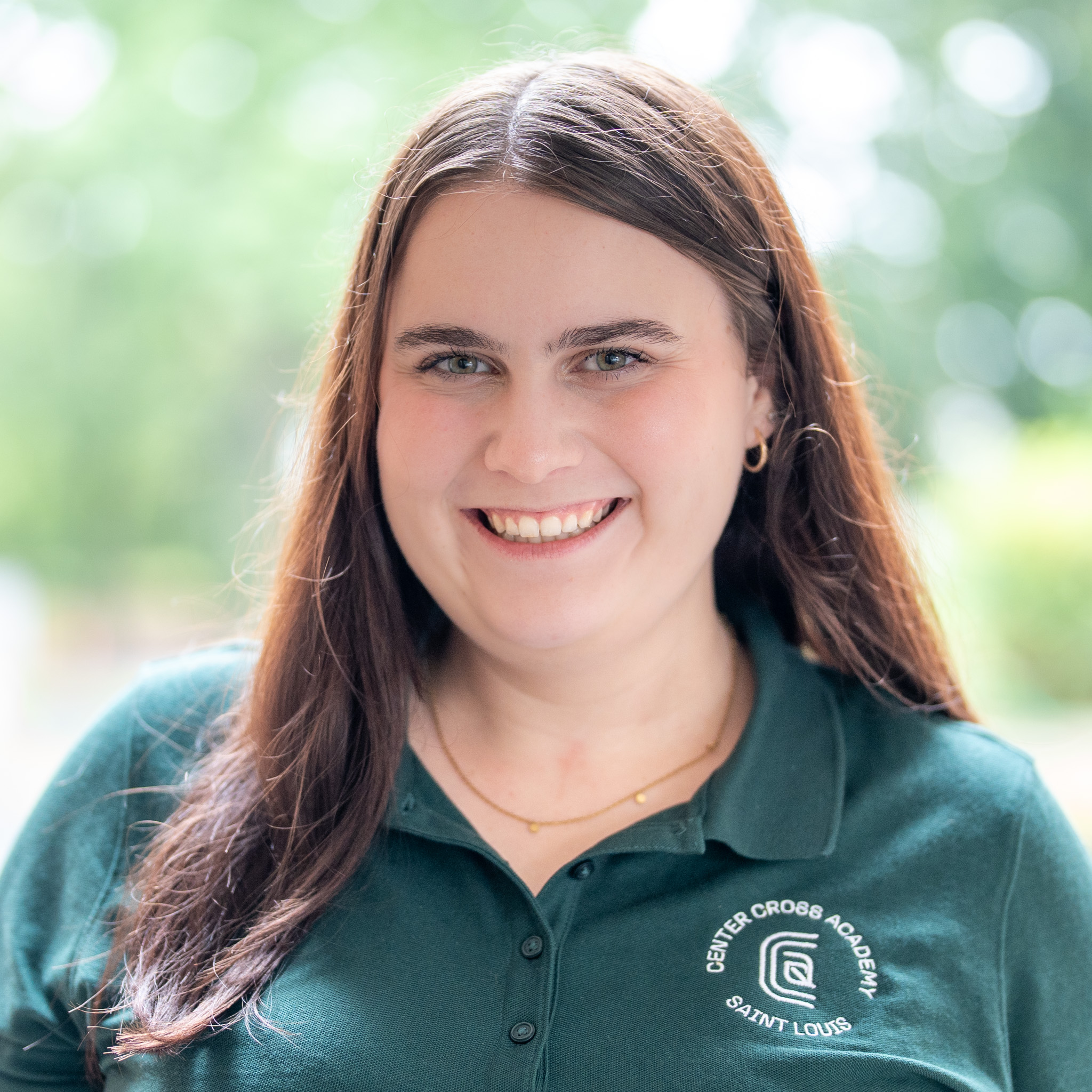 A woman with long brown hair smiles at the camera, wearing a green Center Cross Academy Saint Louis polo shirt. The background is softly blurred greenery.