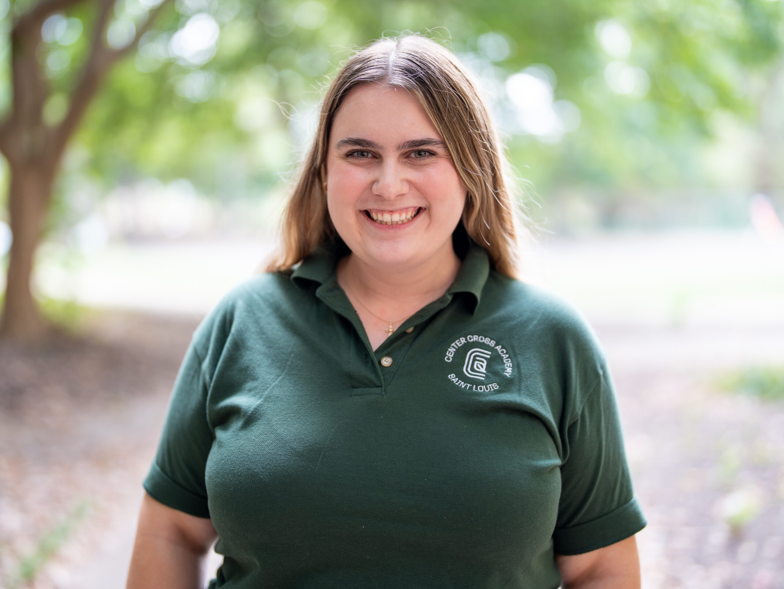A woman with light brown hair smiles outdoors, wearing a dark green polo shirt with the logo and text Green Group School, Saint Louis. She stands in a park-like setting with trees and blurred greenery in the background.