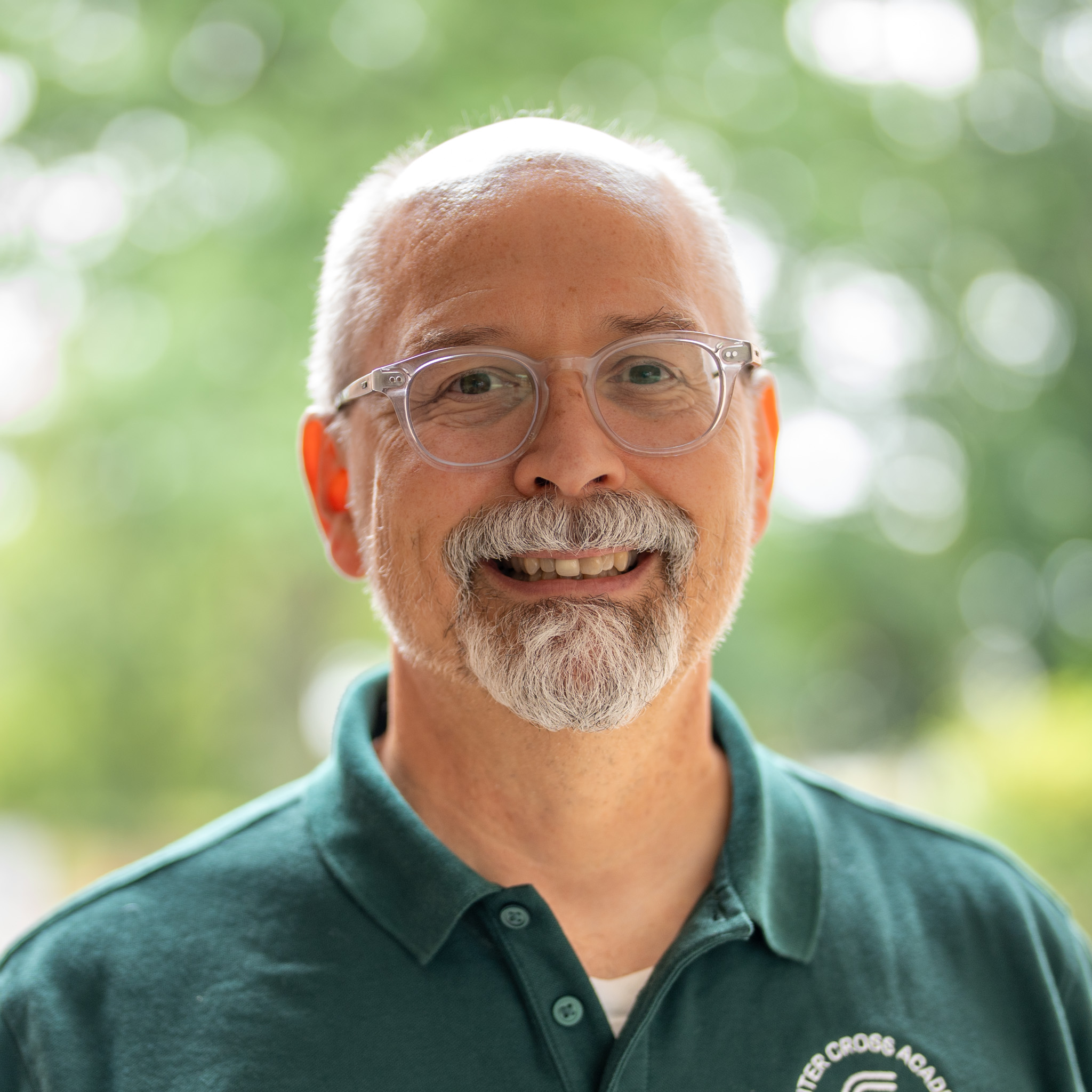 A smiling man with glasses, a white beard, and mustache wearing a green collared shirt stands outdoors with a blurred background of trees and sunlight.