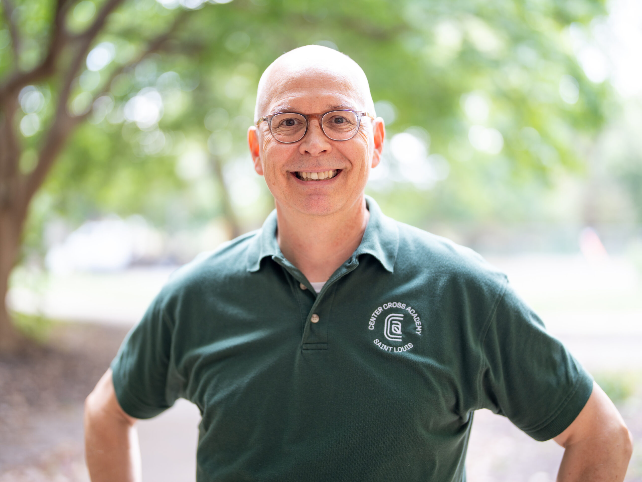 A smiling bald man wearing glasses and a dark green polo shirt stands outdoors with trees in the blurred background. The shirt has a circular white logo on the chest.