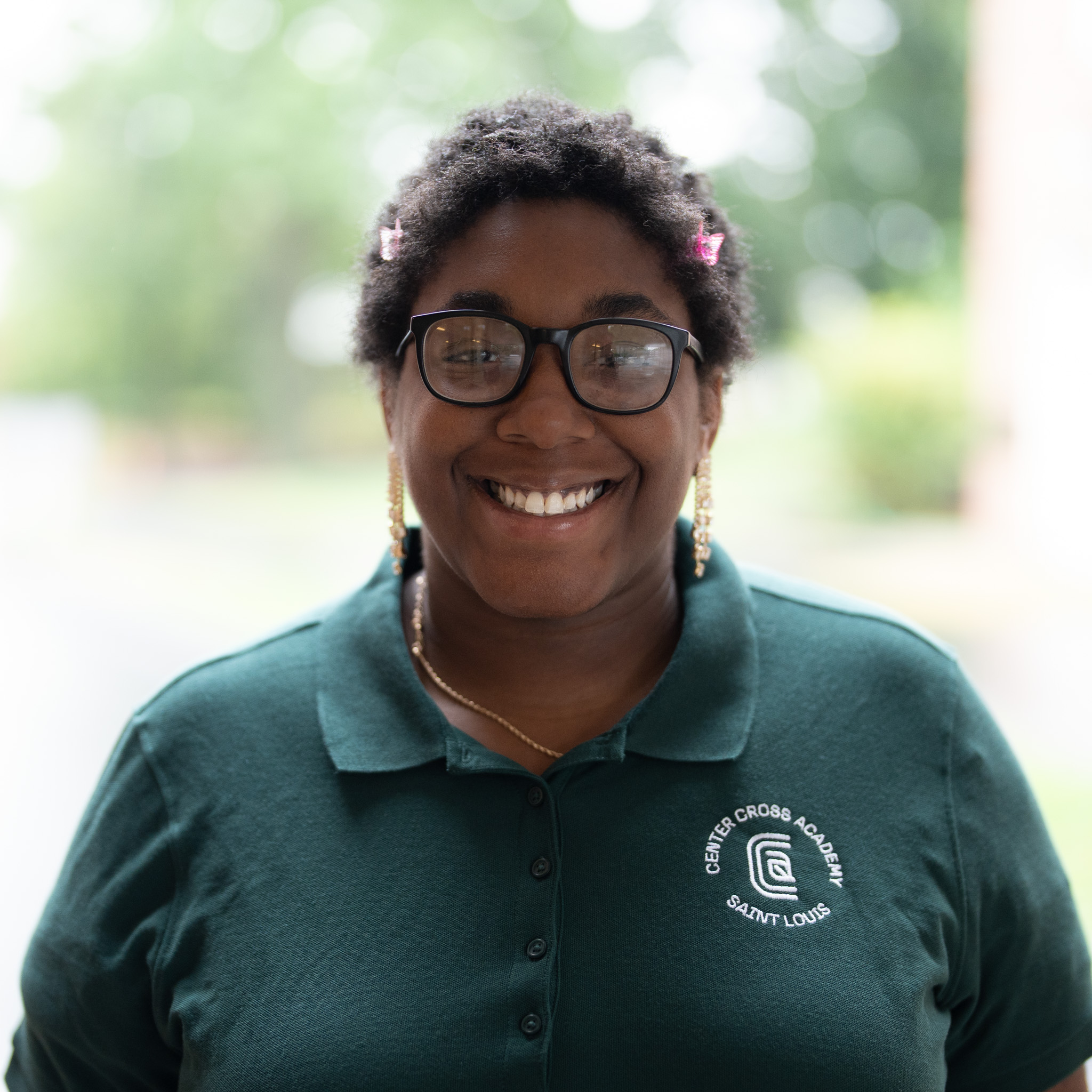 A smiling person with glasses and short curly hair, wearing a green polo shirt with a Genesis Cross Academy St. Louis logo, stands outdoors with a blurred, green background.