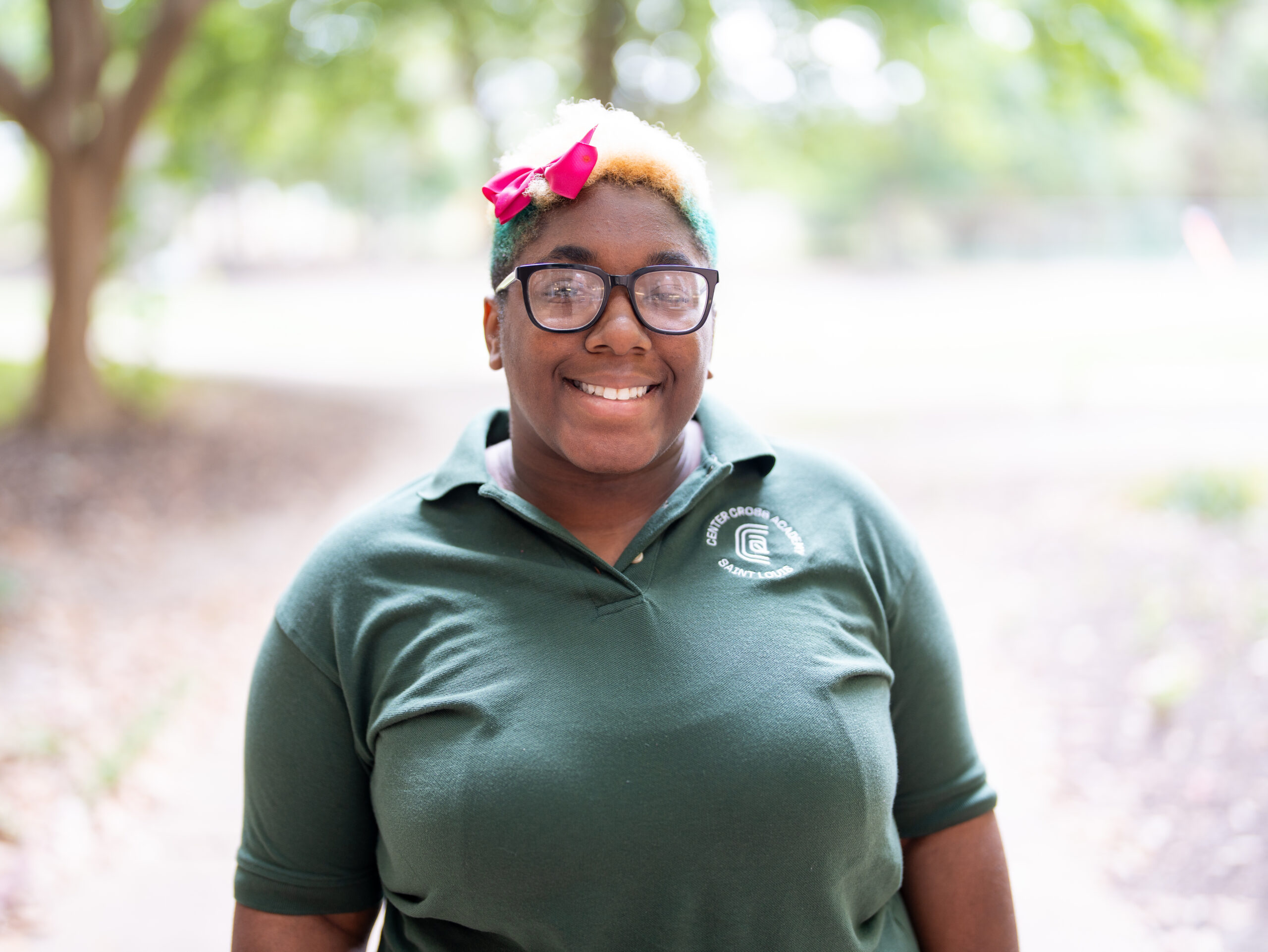 A person wearing glasses, a green polo shirt, and a pink bow in their hair smiles outdoors, with trees and a blurred natural background.