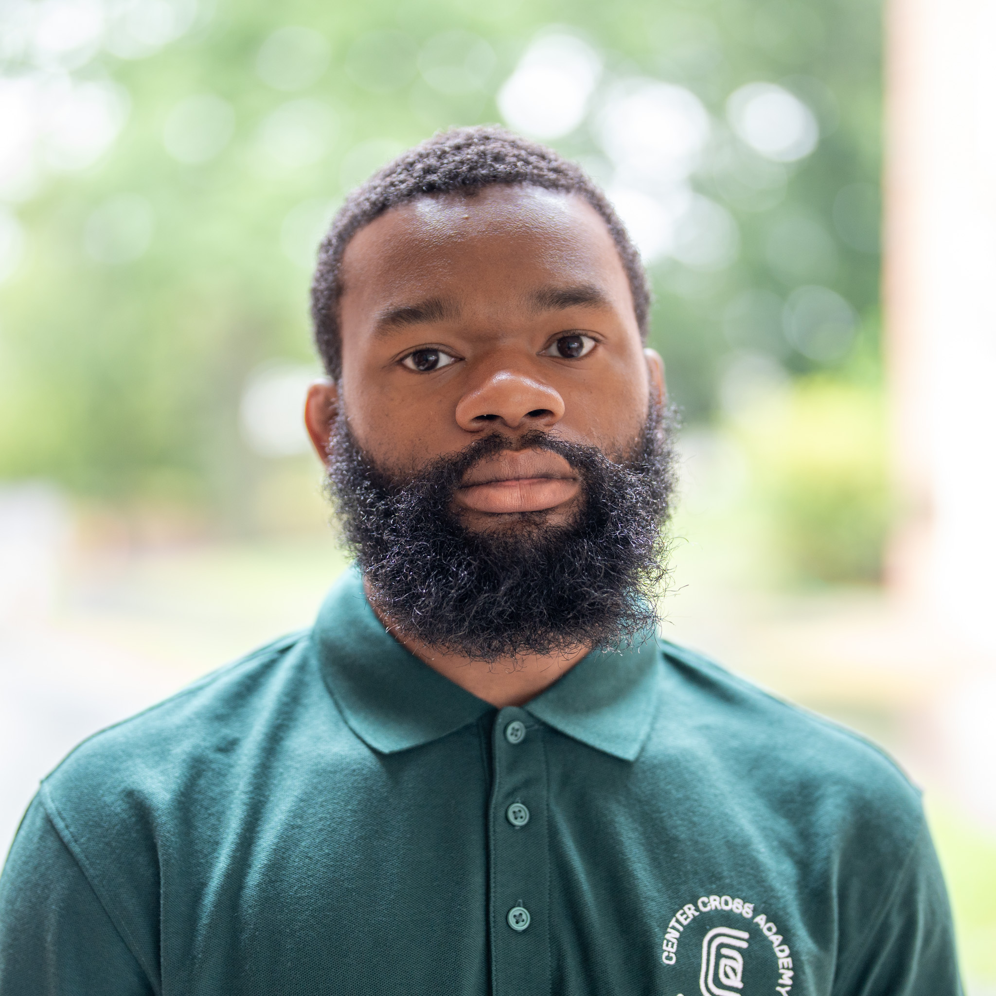 A man with a full beard and short hair wears a green polo shirt with a Center Cross Academy logo, standing outdoors with a blurred, leafy background.