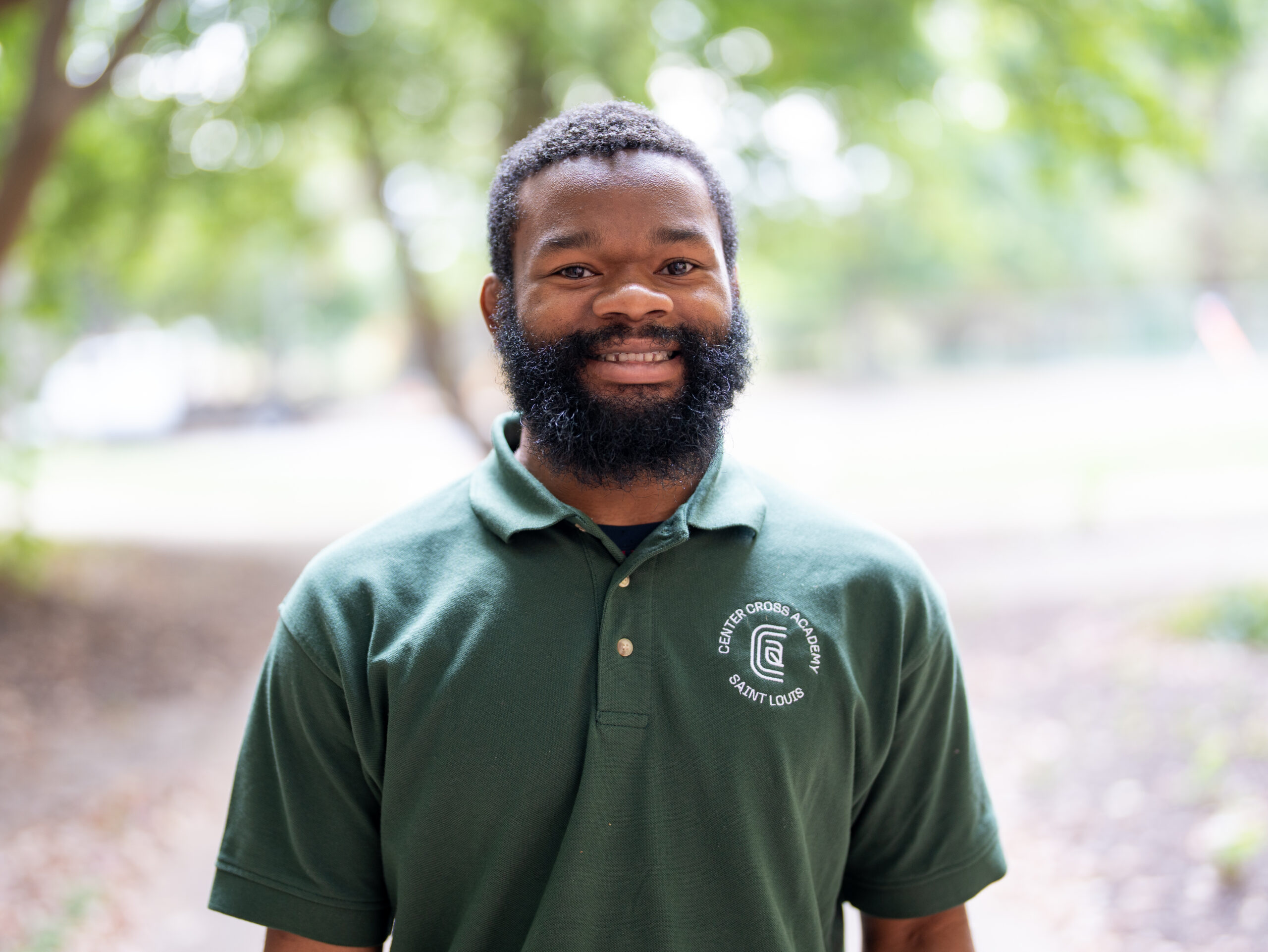 A man with a beard smiles outdoors, wearing a green collared shirt with the logo and text Community Corps Beautify Loop. Trees and foliage are blurred in the background.
