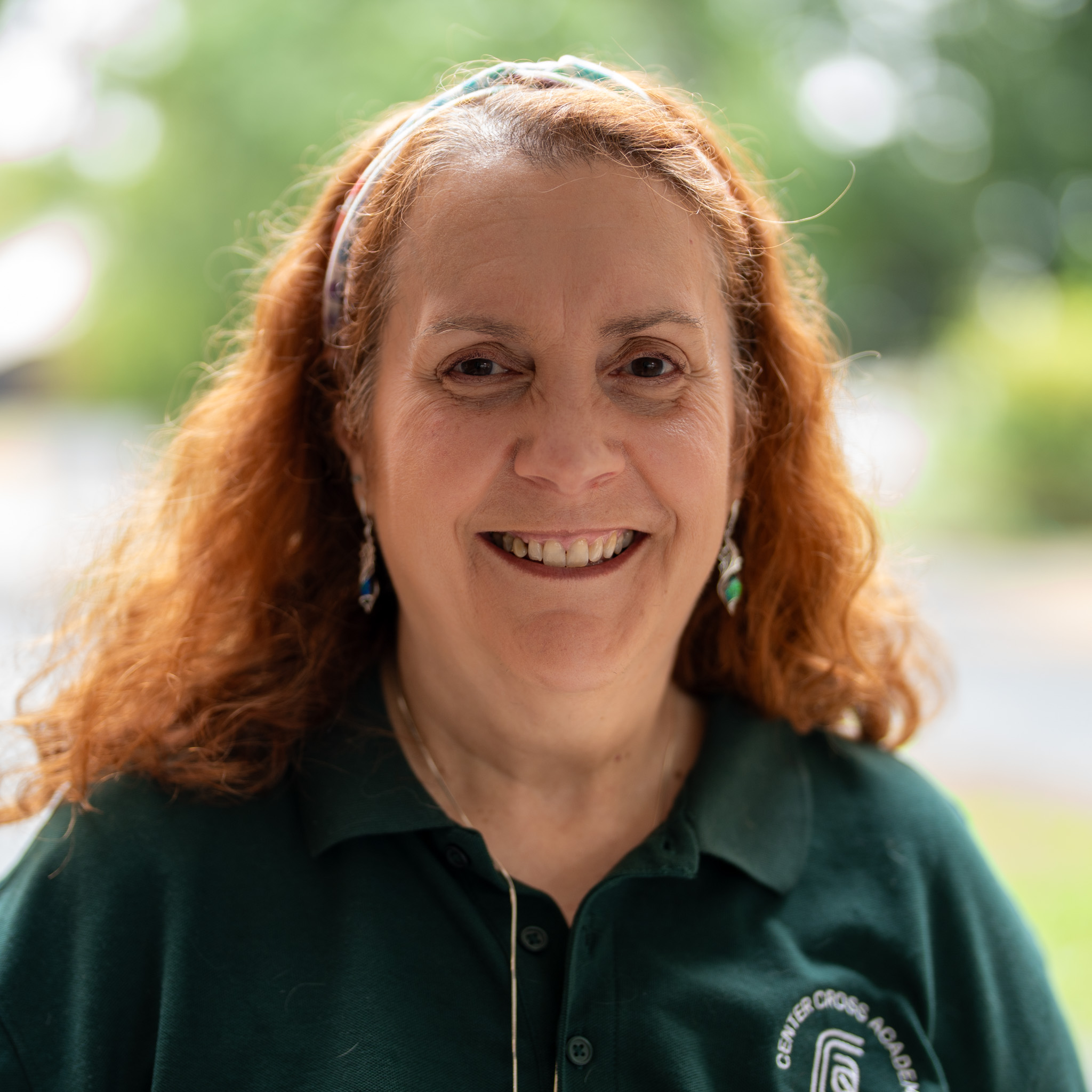 A woman with long red hair, wearing a green collared shirt and a headband, smiles outdoors with blurred greenery in the background.