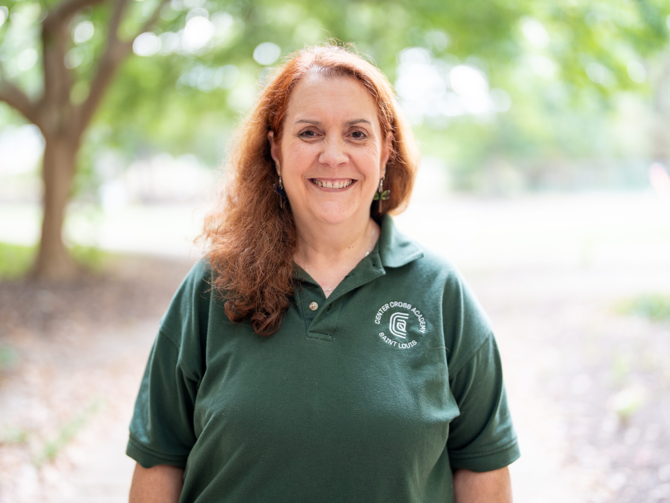 A woman with long red hair smiles outdoors, wearing a dark green polo shirt with a white logo and text on the left side. The background is blurred with greenery and trees.