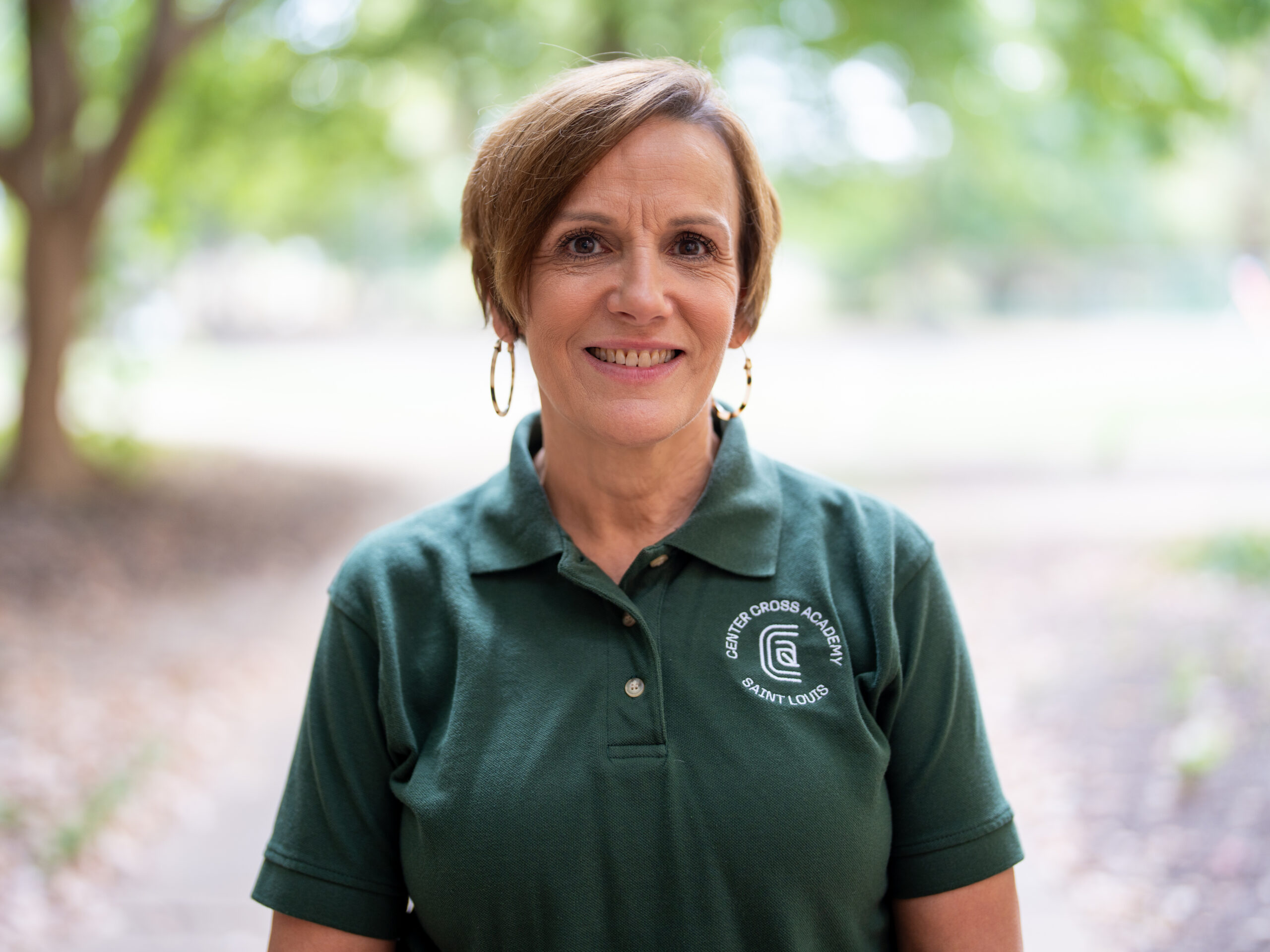 A smiling woman with short brown hair stands outdoors, wearing a green polo shirt with the “Saint George Academy Saint Louis” logo. The background is blurred greenery and trees.