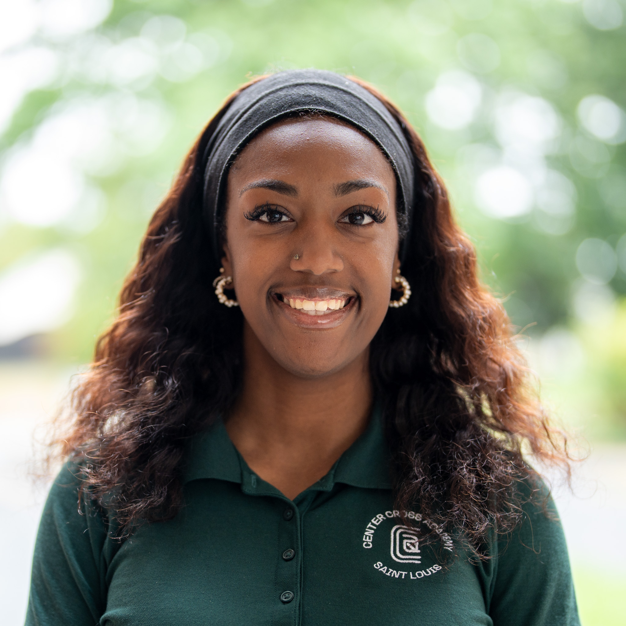 A woman with long, wavy hair and hoop earrings smiles, wearing a dark green collared shirt with a Center for 360° Sports, Saint Louis logo. The background is softly blurred greenery.