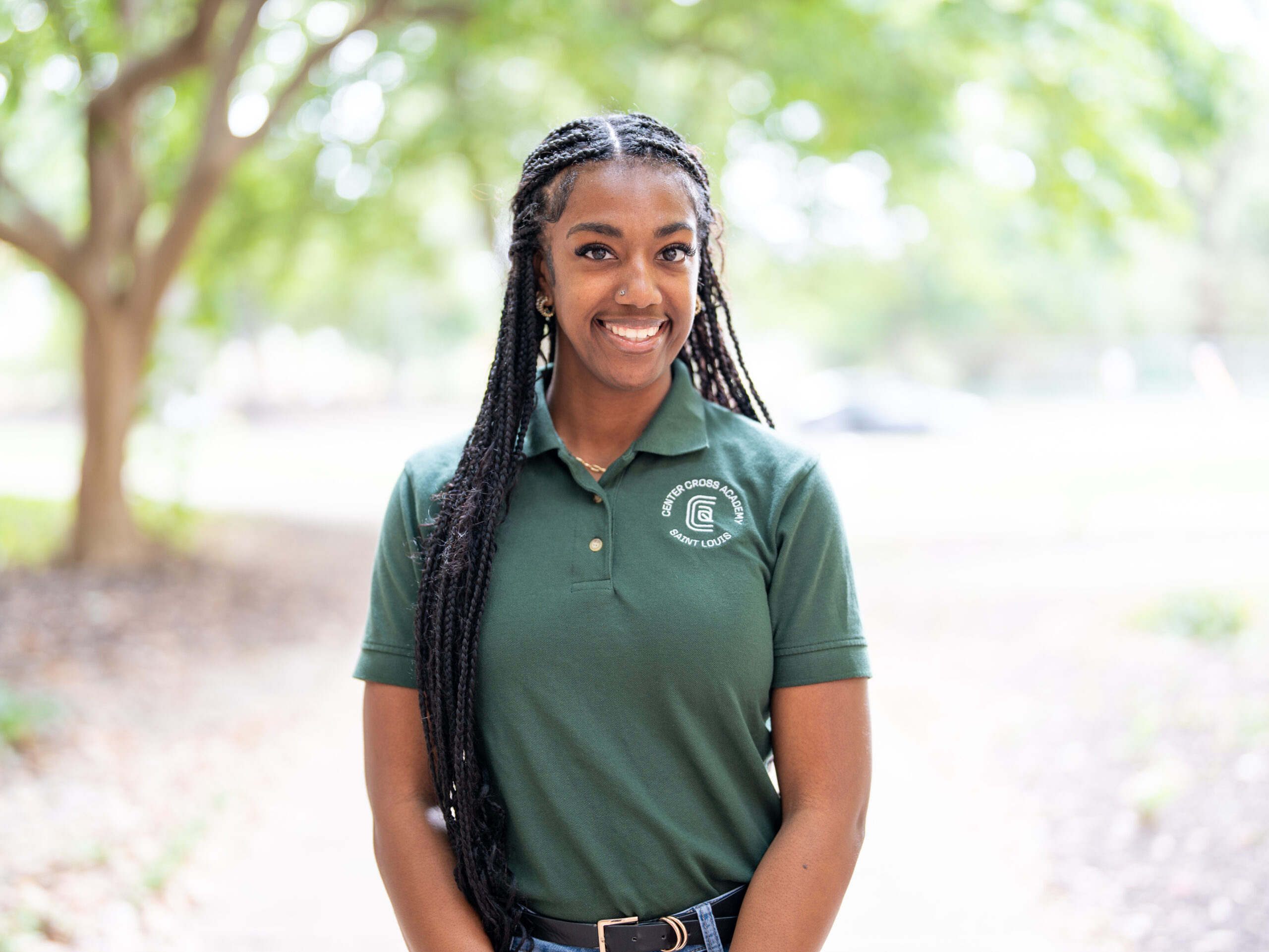 A woman with long braided hair stands outdoors, smiling. She wears a green polo shirt with a logo and blue jeans. The background features trees and soft natural light.