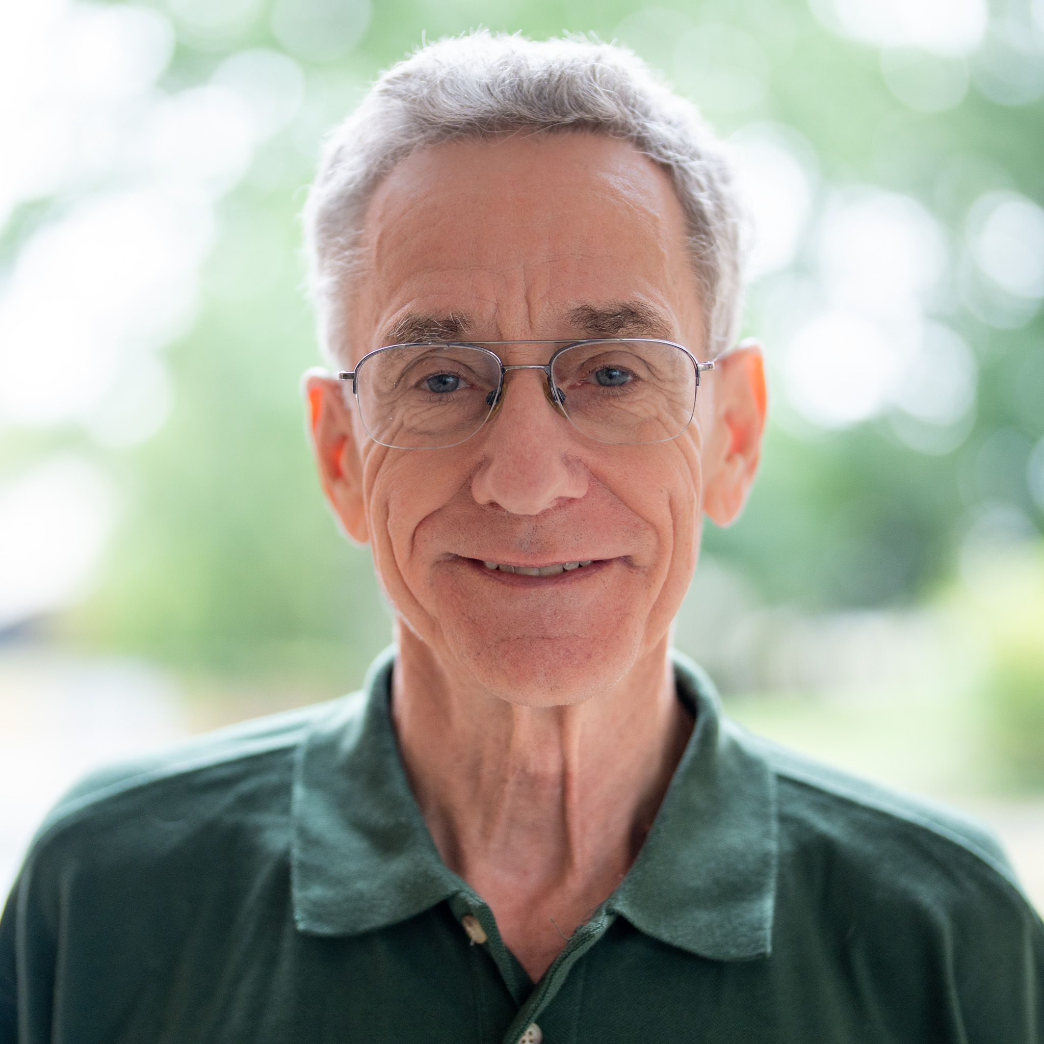 An older man with gray hair and glasses smiles at the camera. He is wearing a dark green polo shirt and standing outdoors with a blurred green background.