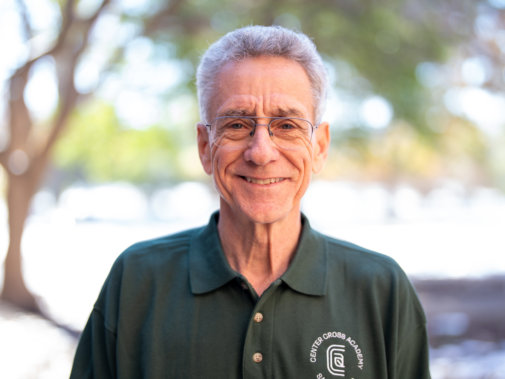 An older man with gray hair and glasses smiles outdoors. He is wearing a dark green polo shirt with the CENTER CROSS ACADEMY logo. The background is blurred with green and yellow trees.