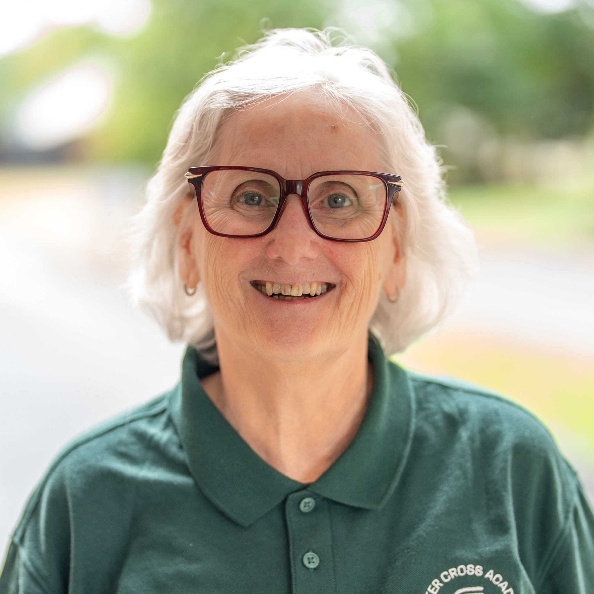 Smiling older woman with short white hair and large glasses, wearing a green collared shirt, standing outdoors with a blurred background of trees and sunlight.