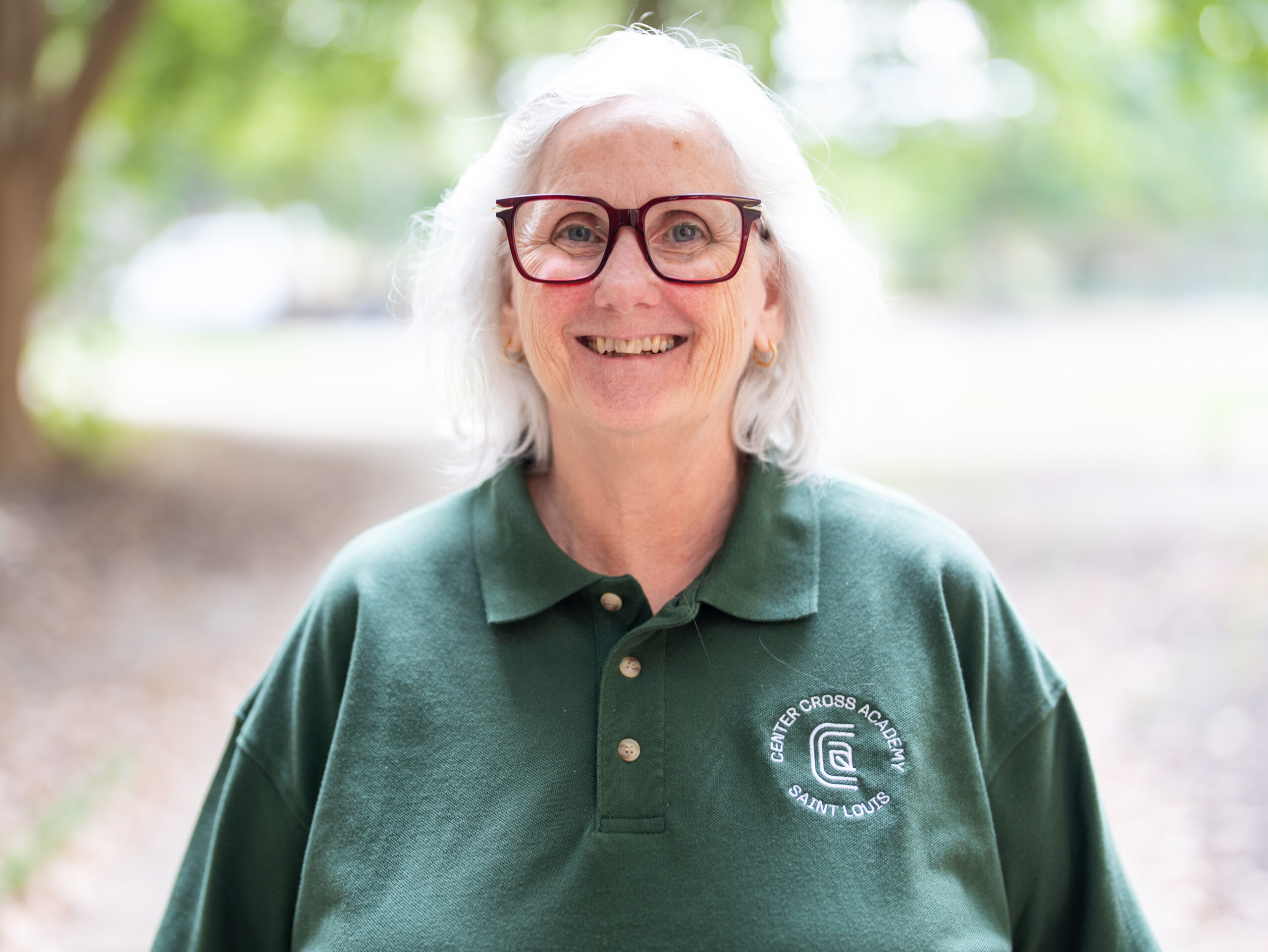 An older woman with white hair and glasses smiles outdoors. She is wearing a green polo shirt with a Center Cross Academy Saint Louis logo. The background is blurred greenery.