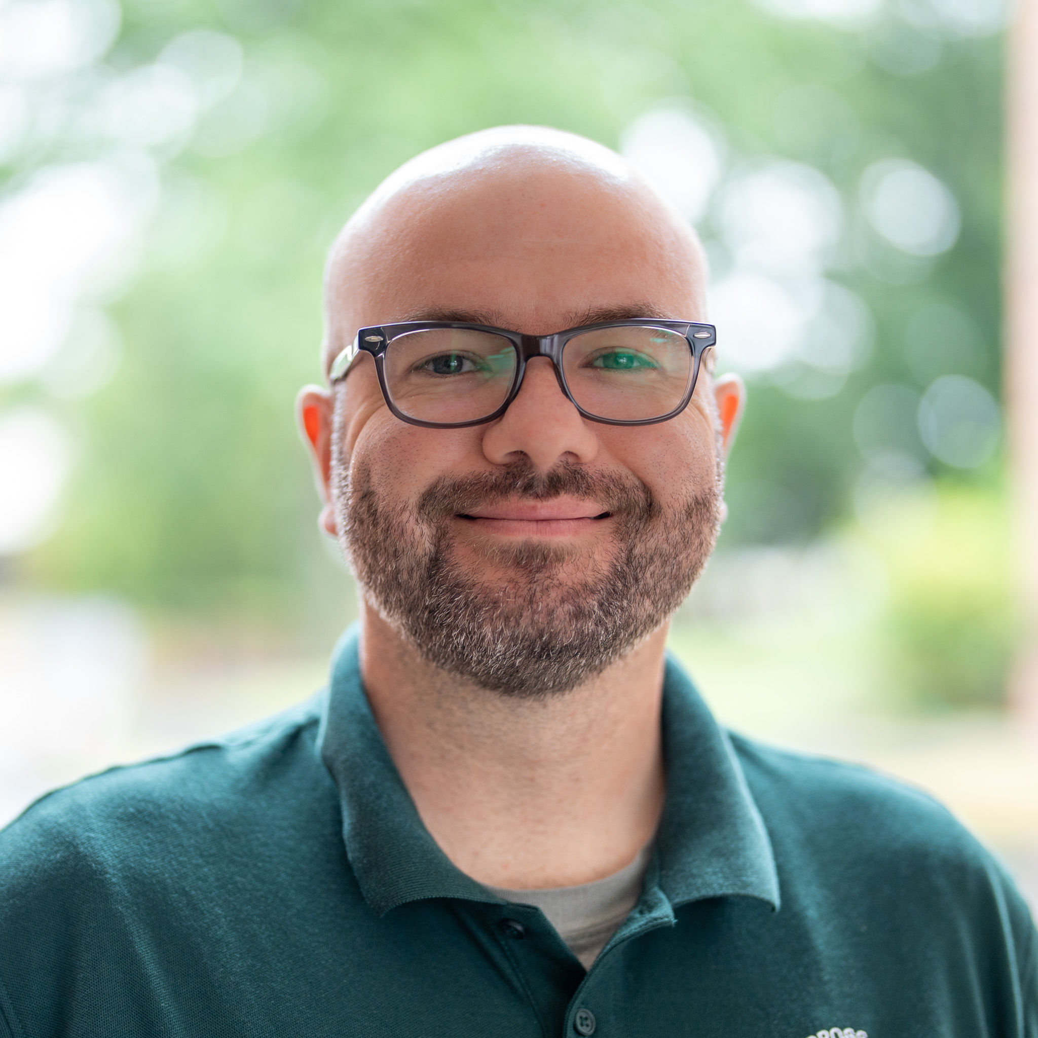 A smiling man with glasses, a beard, and a bald head wears a green collared shirt. The background is blurred with greenery and natural light.