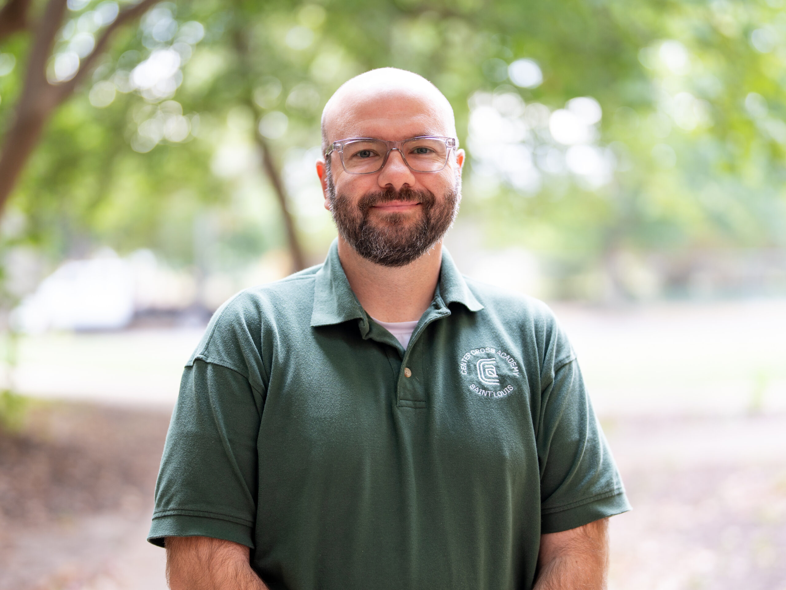 A man with glasses, a beard, and a shaved head stands outdoors, smiling. He wears a green collared shirt with a white logo on the left side. The background is blurred with green trees and sunlight.