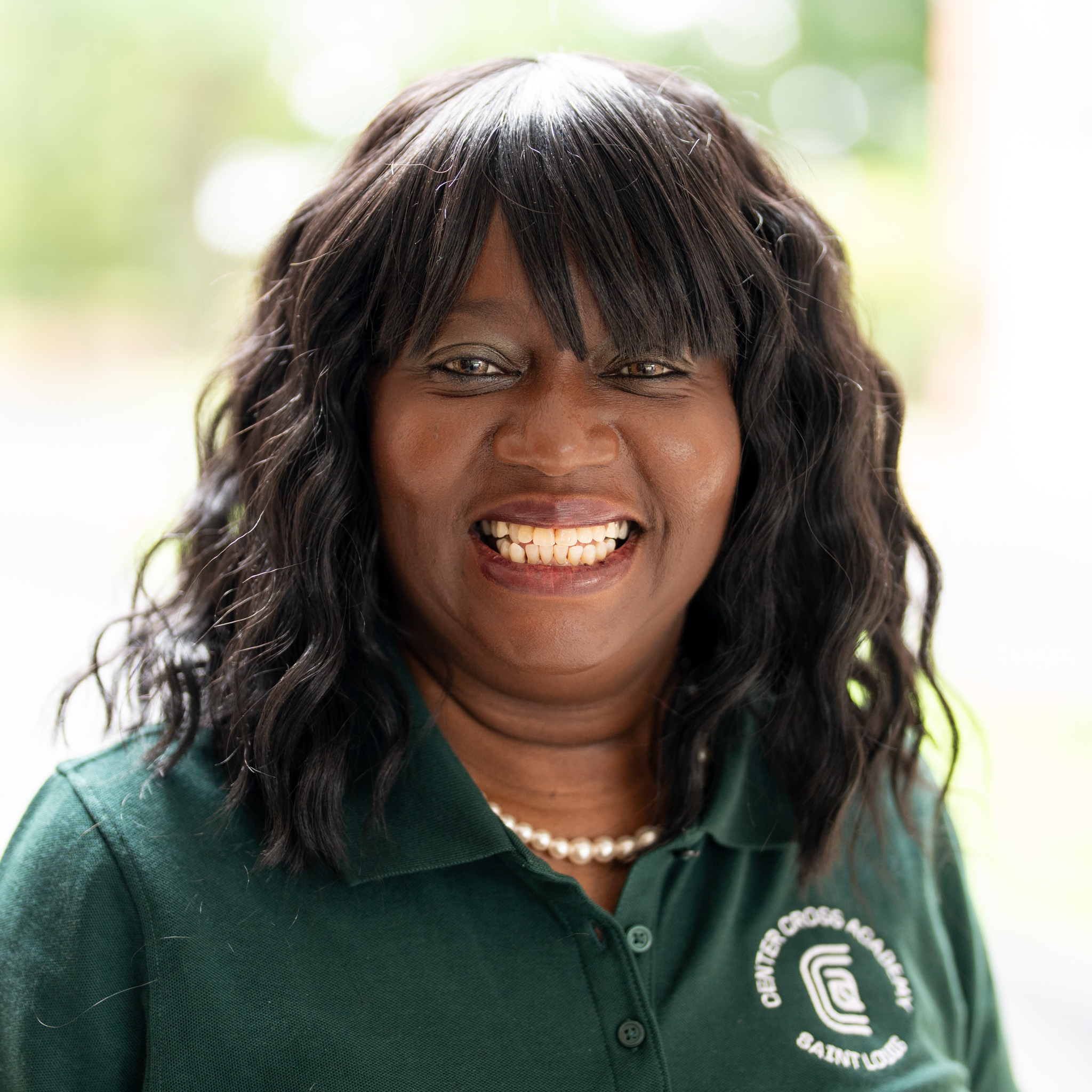 Smiling woman with long wavy hair wearing a green polo shirt with a logo and pearls, standing outdoors with a blurred background.