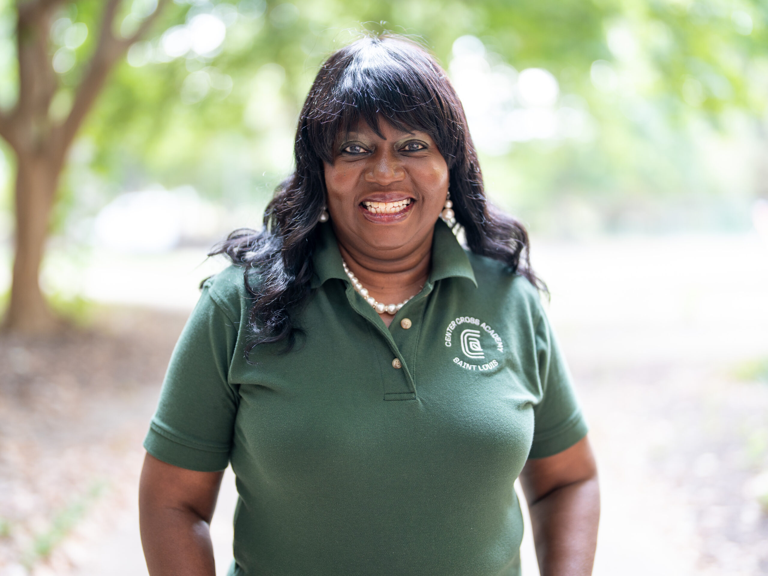 A woman with long dark hair smiles outdoors, wearing a dark green polo shirt with a logo and pearl necklace. The background is blurred with green trees and sunlight.