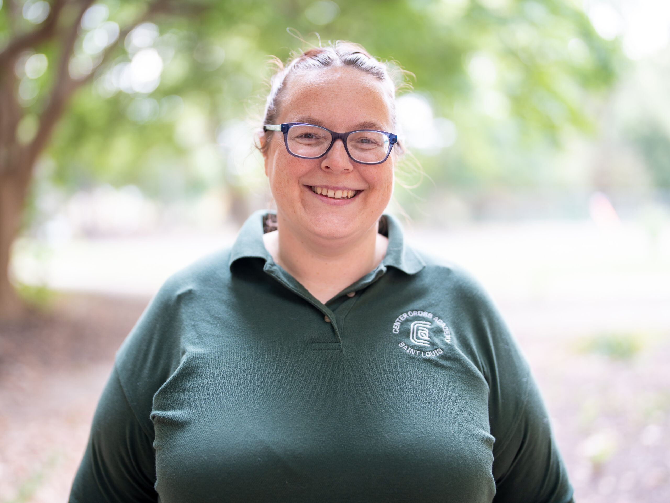 A smiling person with glasses and a green collared shirt stands outdoors with trees and greenery in the blurred background.