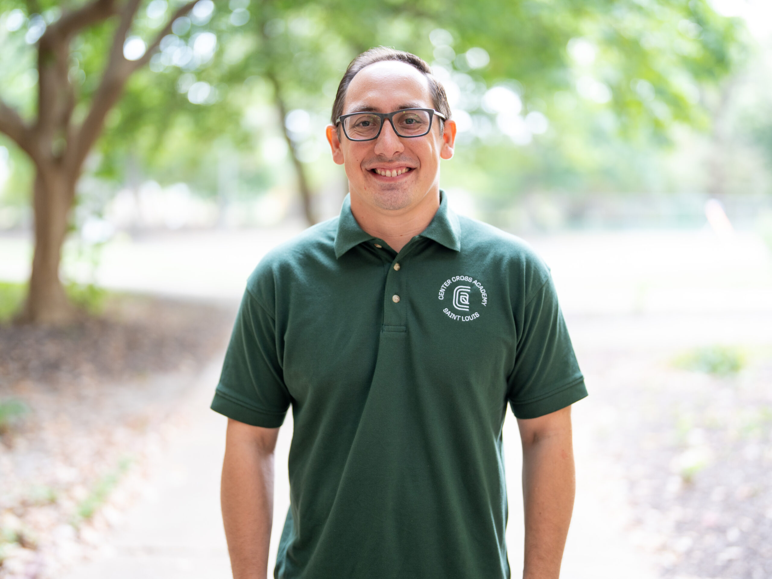 A person wearing glasses and a dark green polo shirt with a circular white logo stands outdoors on a pathway, smiling, with blurred trees and greenery in the background.