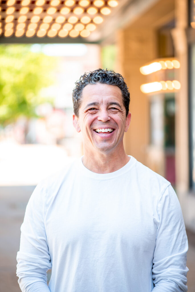 A man with short curly hair, wearing a white long-sleeve shirt, stands outside and smiles broadly at the camera. The background is softly blurred with lights and greenery visible.