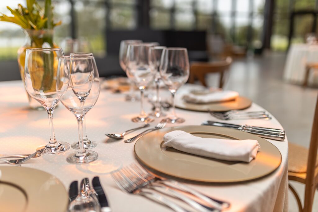 A close-up of an elegant table setting with gold-rimmed plates, neatly folded white napkins, multiple wine glasses, and silver cutlery, set in a bright, modern dining area.