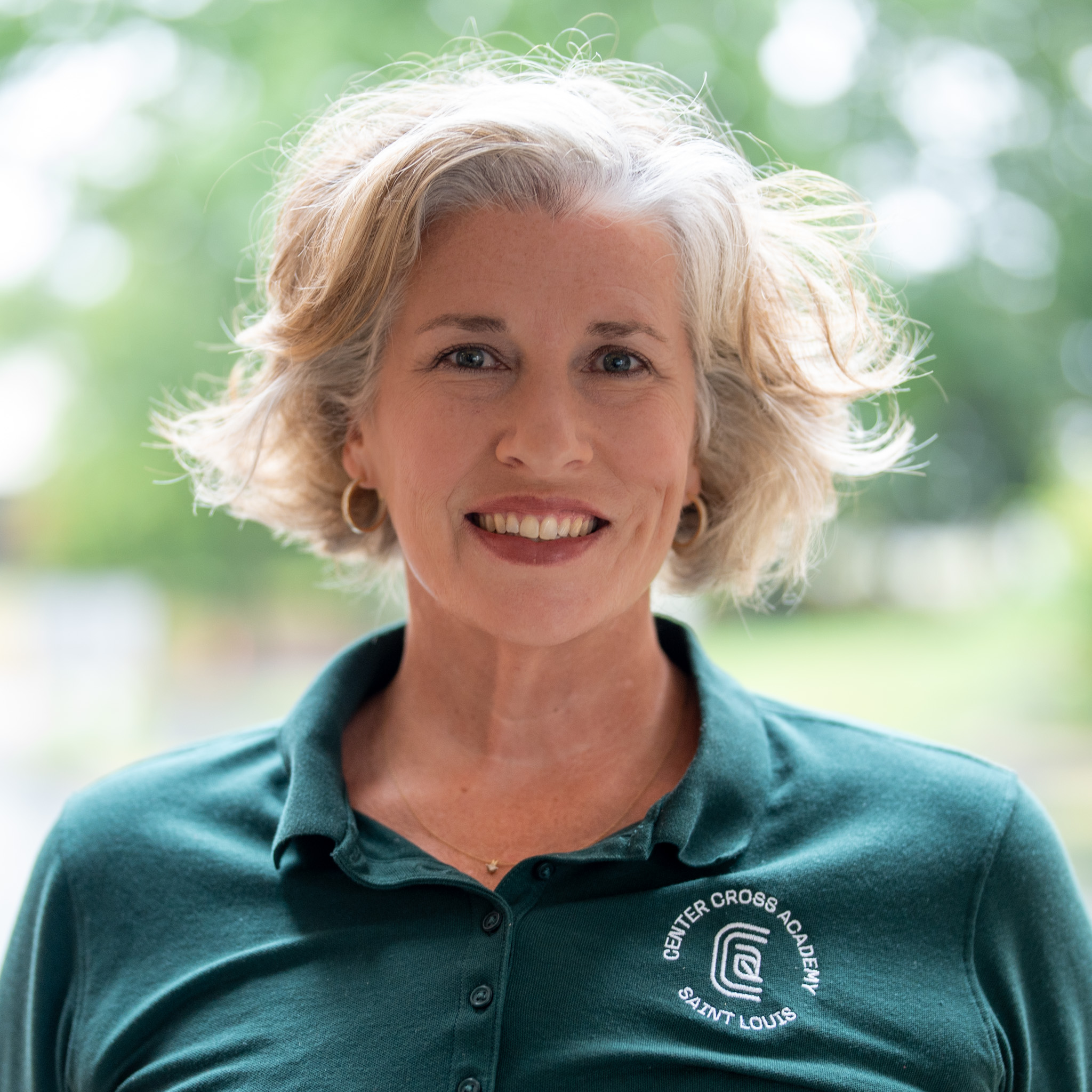 Smiling woman with short gray hair wearing a green Center Cross Academy Saint Louis polo shirt, standing outdoors with a blurred green background.