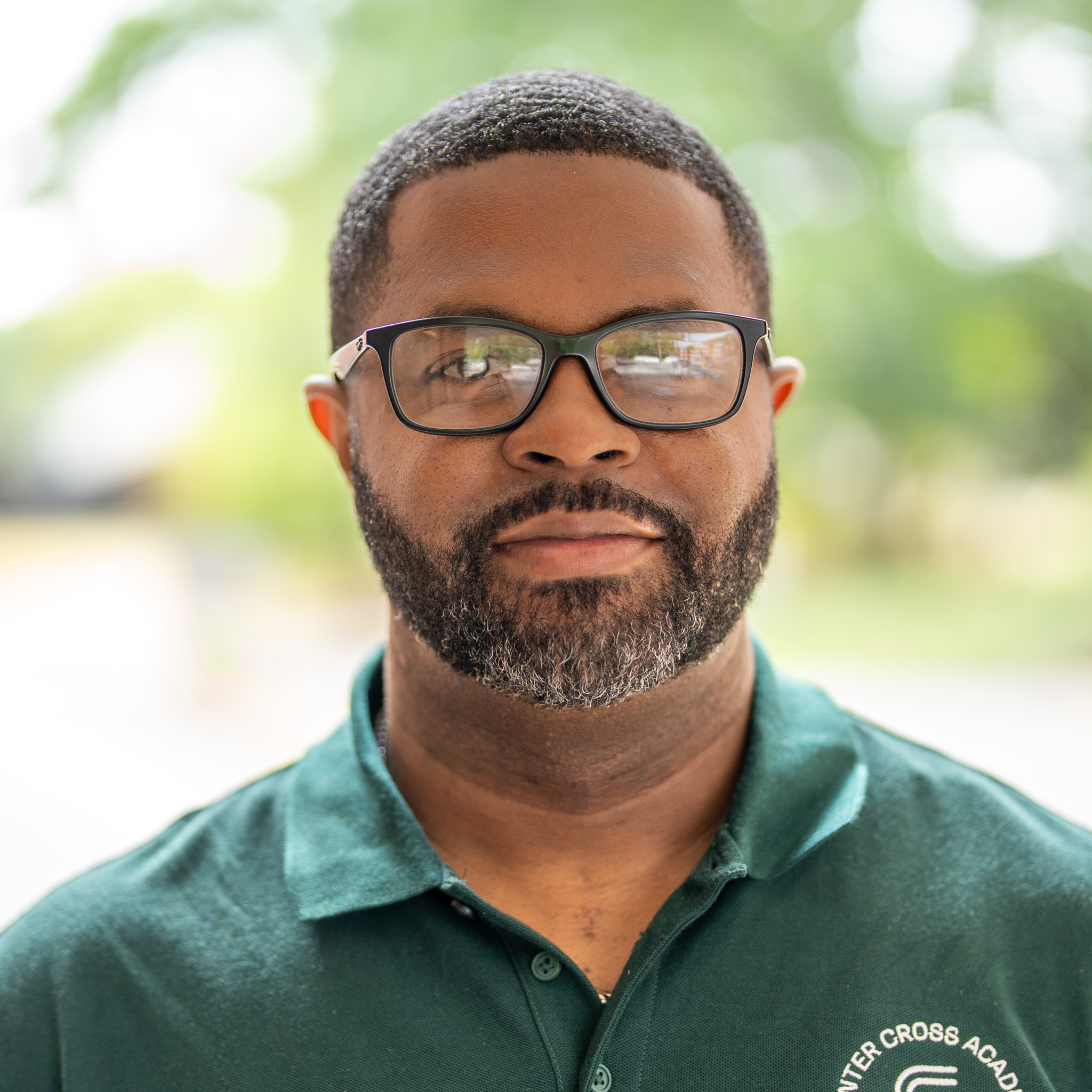 A man with short hair, glasses, and a trimmed beard wearing a green collared shirt stands outside with a blurred green background.