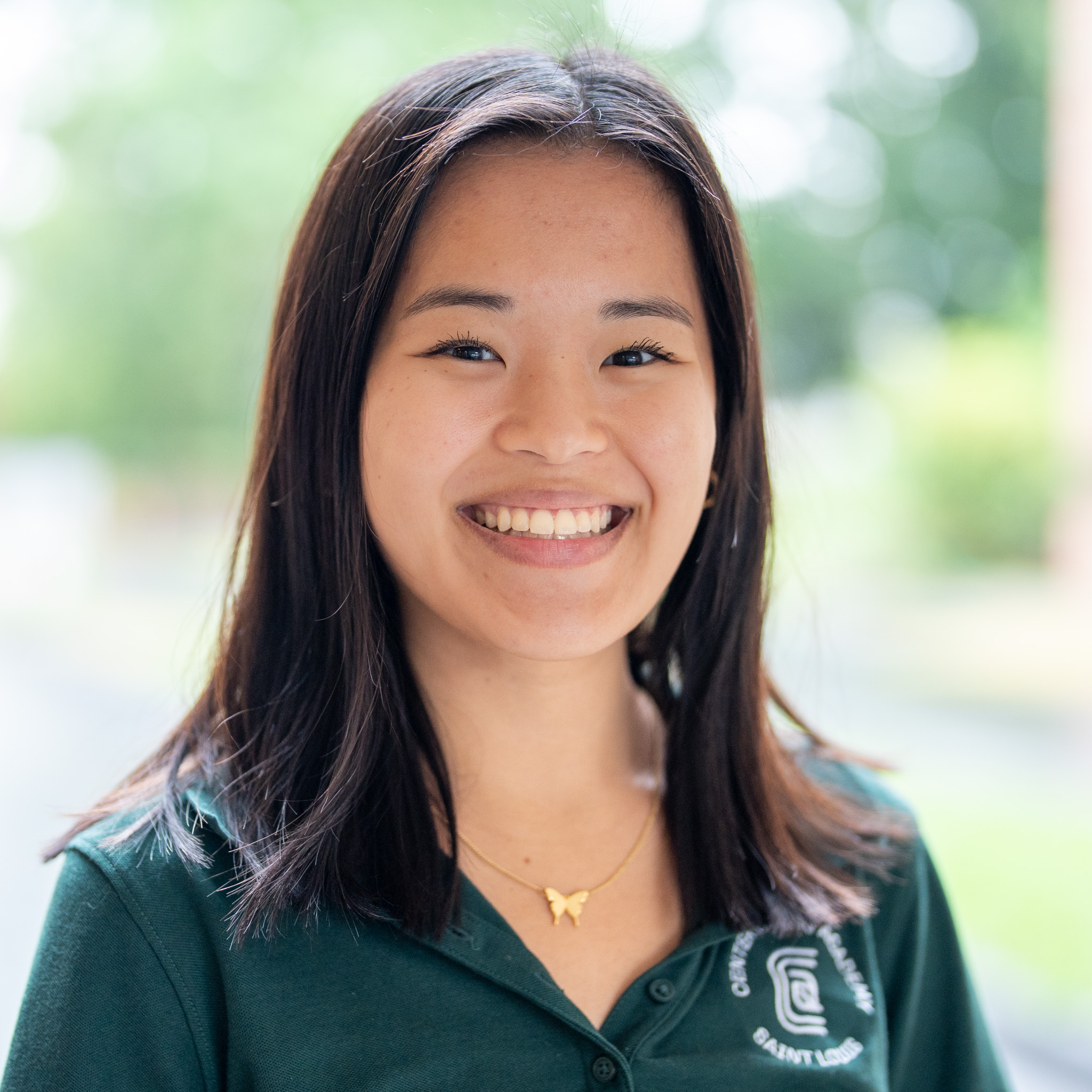 A young woman with straight dark hair smiles outdoors, wearing a dark green collared shirt with a white logo and a butterfly necklace.