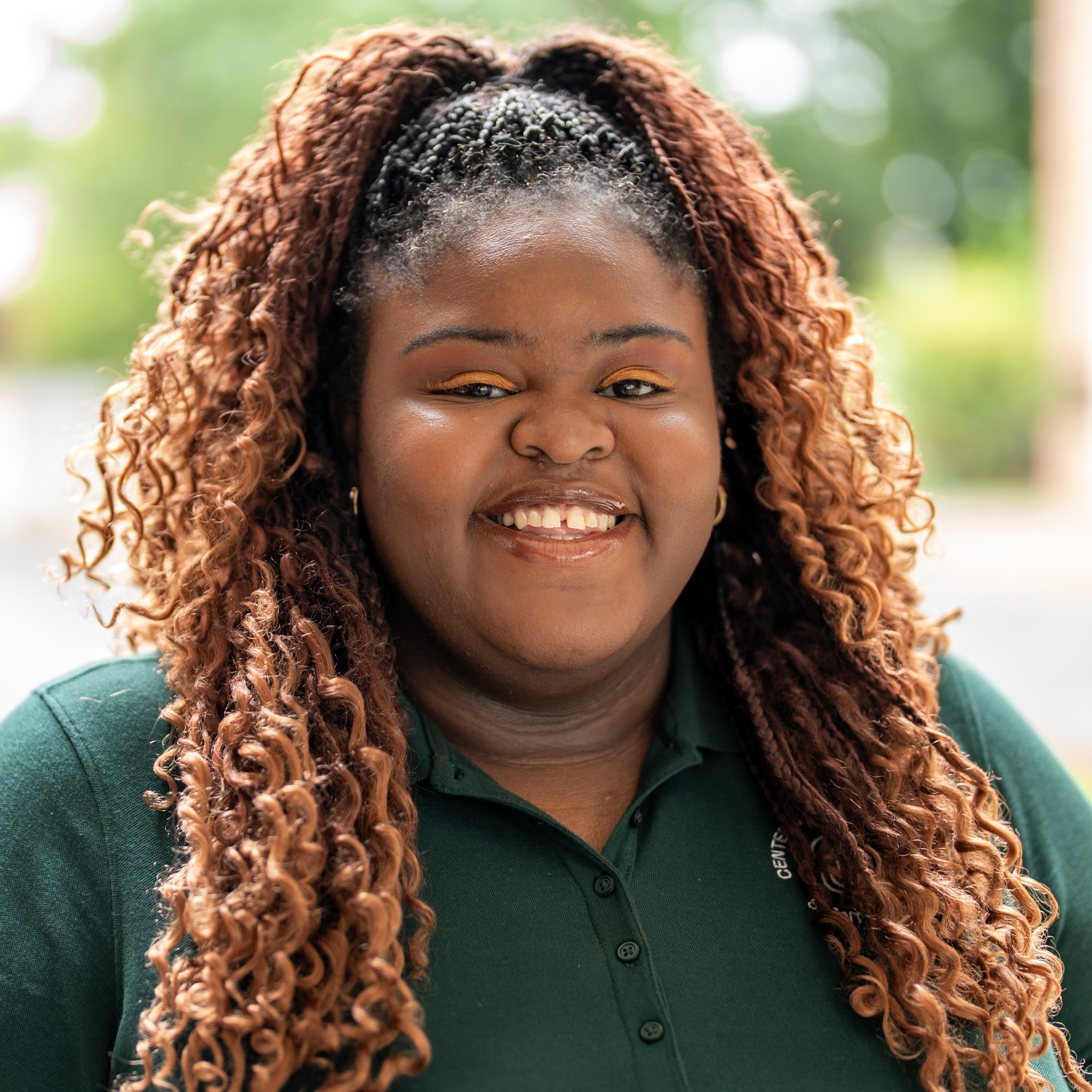 A woman with long, curly brown hair and gold eyeshadow smiles outdoors, wearing a dark green collared shirt. Trees and greenery are blurred in the background.