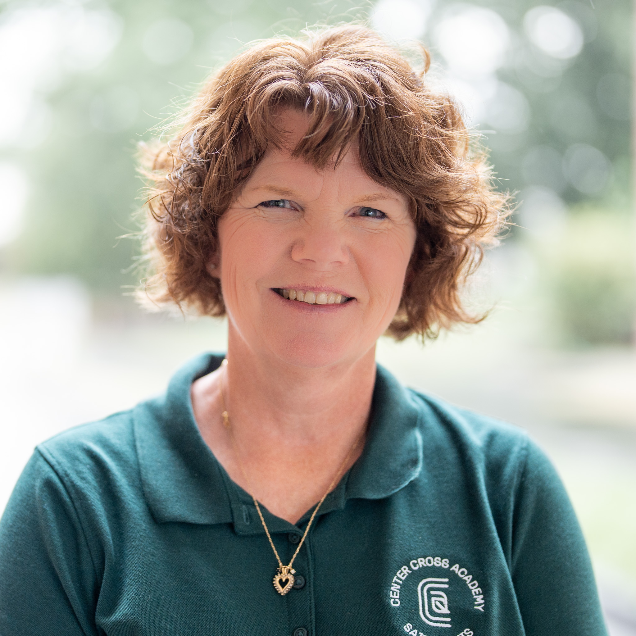A woman with curly brown hair smiles, wearing a green polo shirt with a Center Cross Academy logo and a heart necklace, standing outdoors with a blurred background.